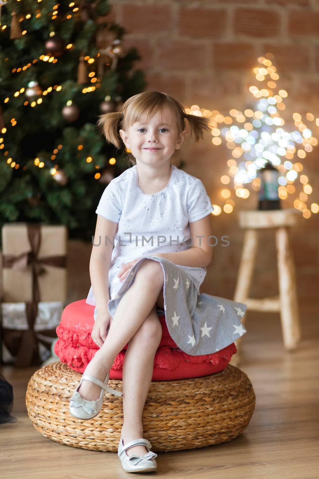 Pretty little child is sitting in front of christmas tree among garlands and presents..