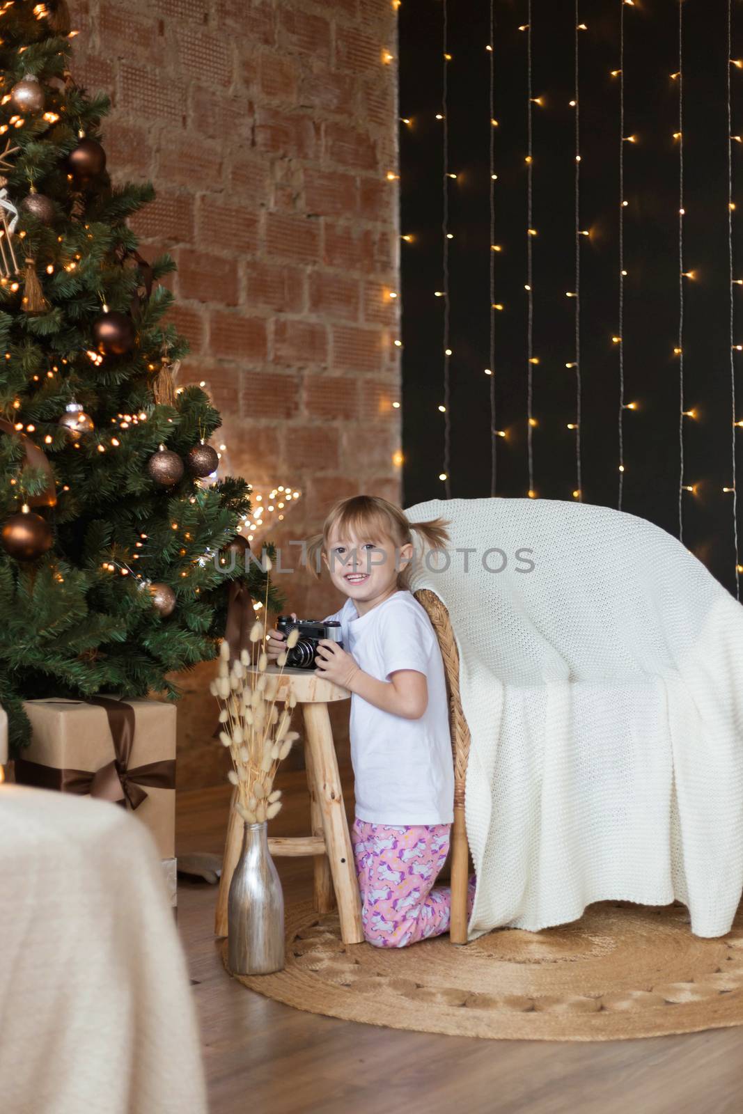 Adorable little girl in pajamas sits on the floor with camera in her hands near christmas tree.