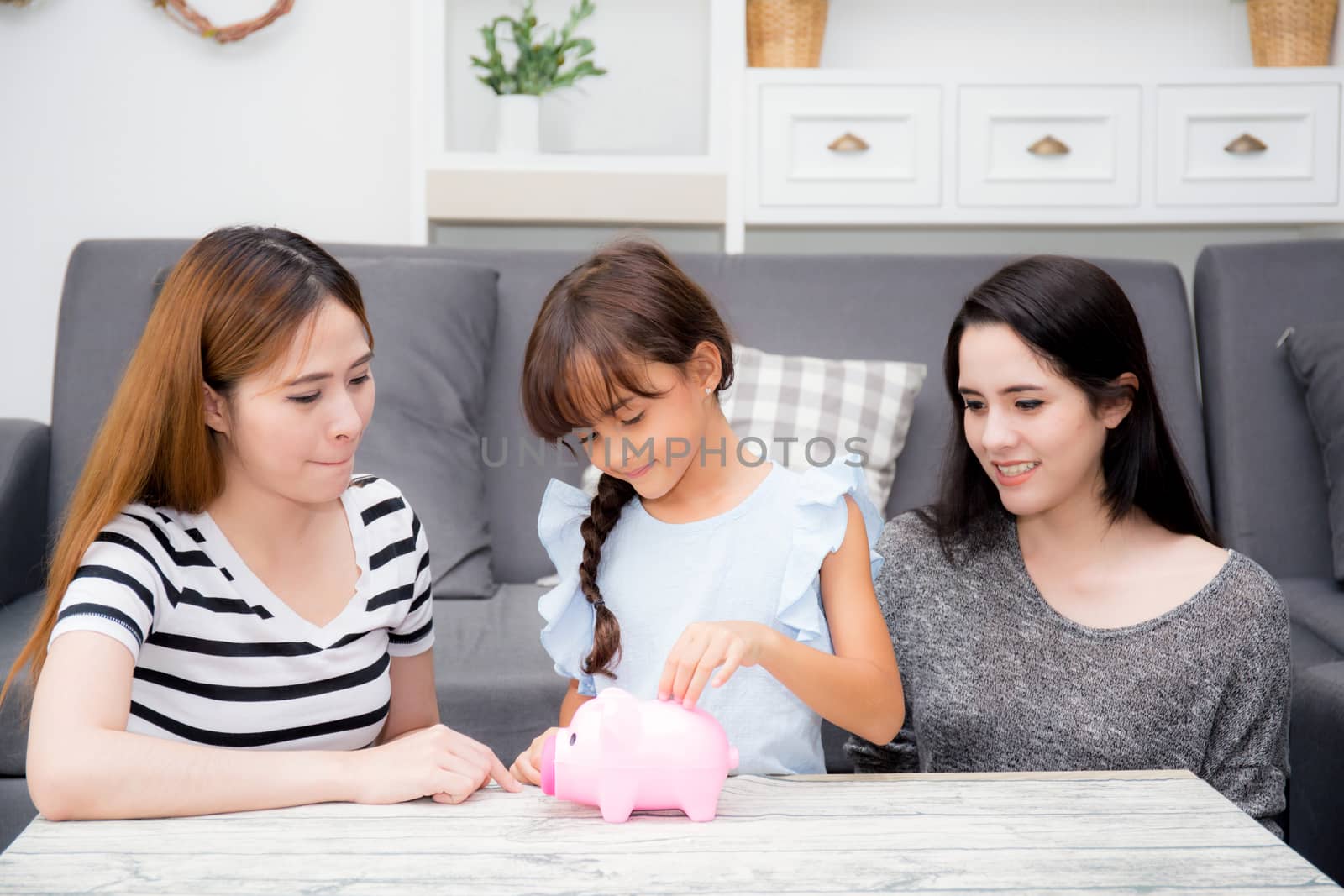Asian mother and aunt smile and happy see daughter putting coin in piggy bank for saving in the living room at home, kid put money with deposit banking for future, family concept.