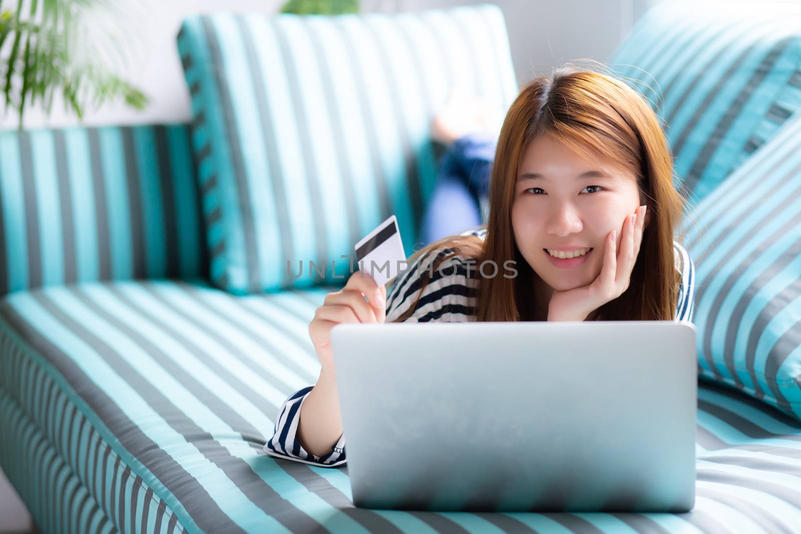 Beautiful of portrait young asian woman lying users credit card with laptop, Content girl shopping online and payment with notebook computer on sofa, lifestyle concept.