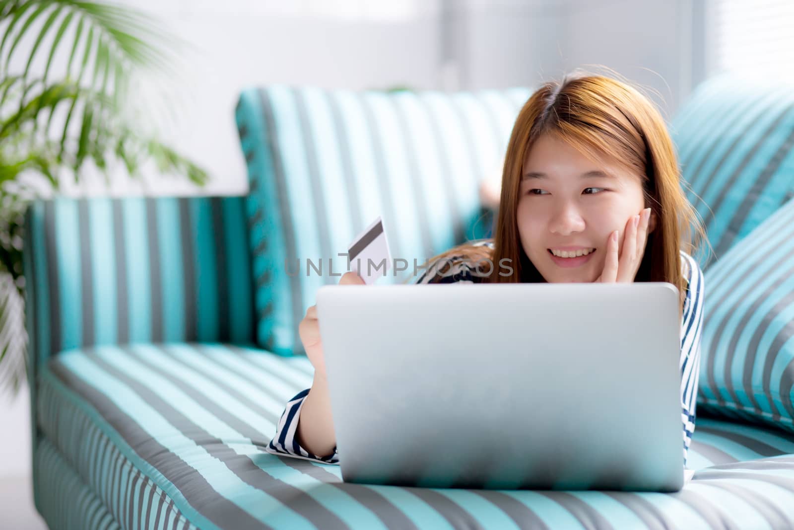 Beautiful of portrait young asian woman lying users credit card with laptop, Content girl shopping online and payment with notebook computer on sofa, lifestyle concept.