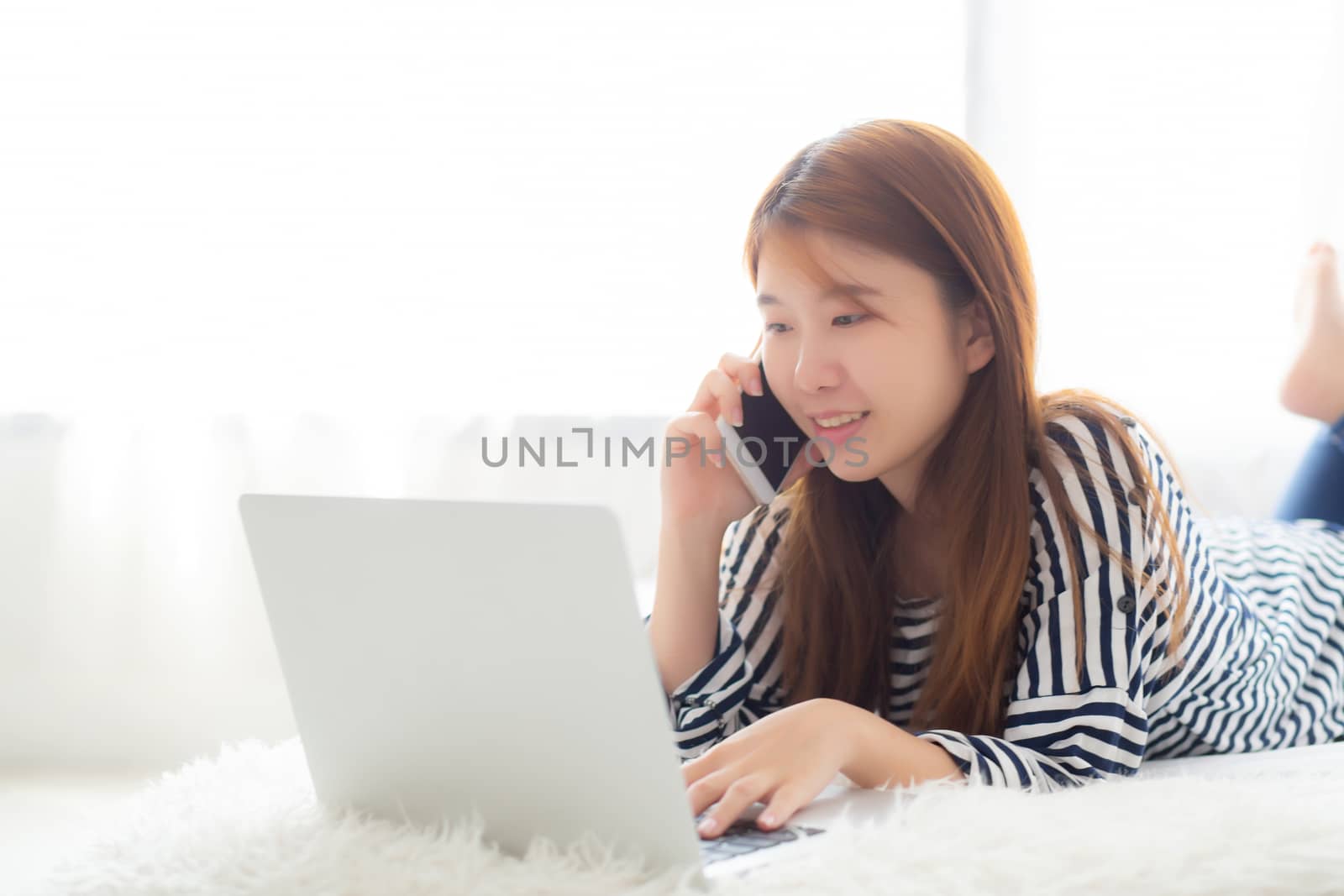Beautiful asian young woman lying on bed using laptop and talking mobile smart phone at bedroom for leisure and relax, freelance with girl working notebook, communication concept.