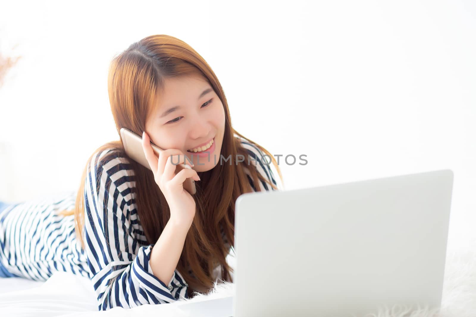 Beautiful asian young woman lying on bed using laptop and talking mobile smart phone at bedroom for leisure and relax, freelance with girl working notebook, communication concept.