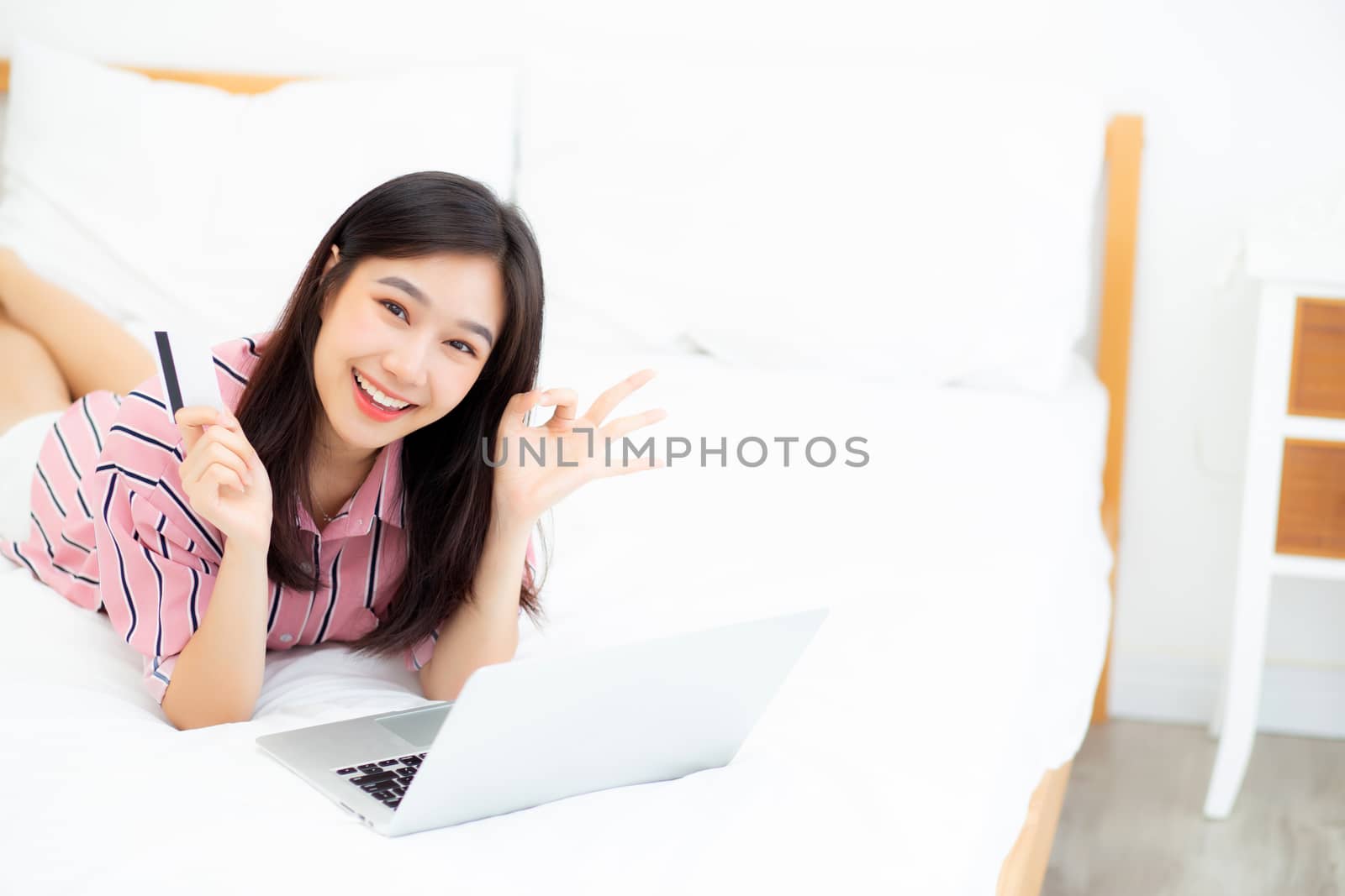 Beautiful of portrait young asian woman lying users credit card with laptop, content girl shopping online and payment with notebook computer on bed at bedroom, lifestyle concept.