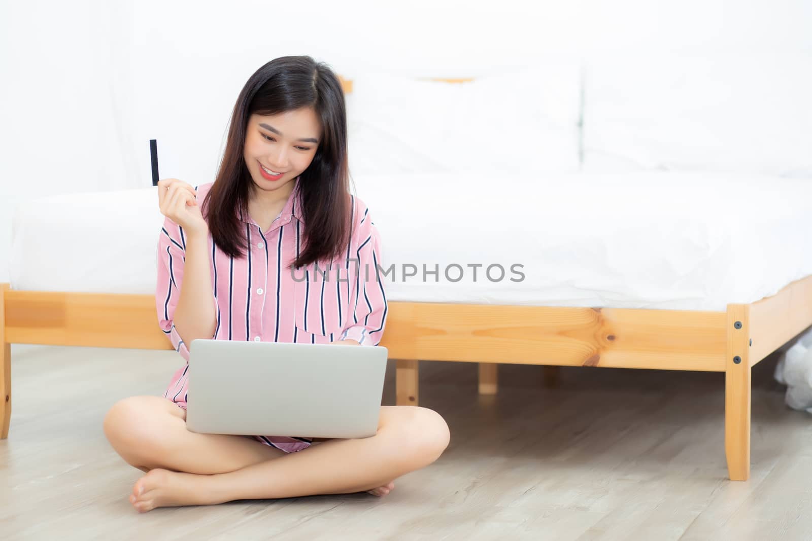 Beautiful of portrait young asian woman sitting users credit card with laptop, content girl shopping online and payment with notebook computer on bed at bedroom, lifestyle concept.