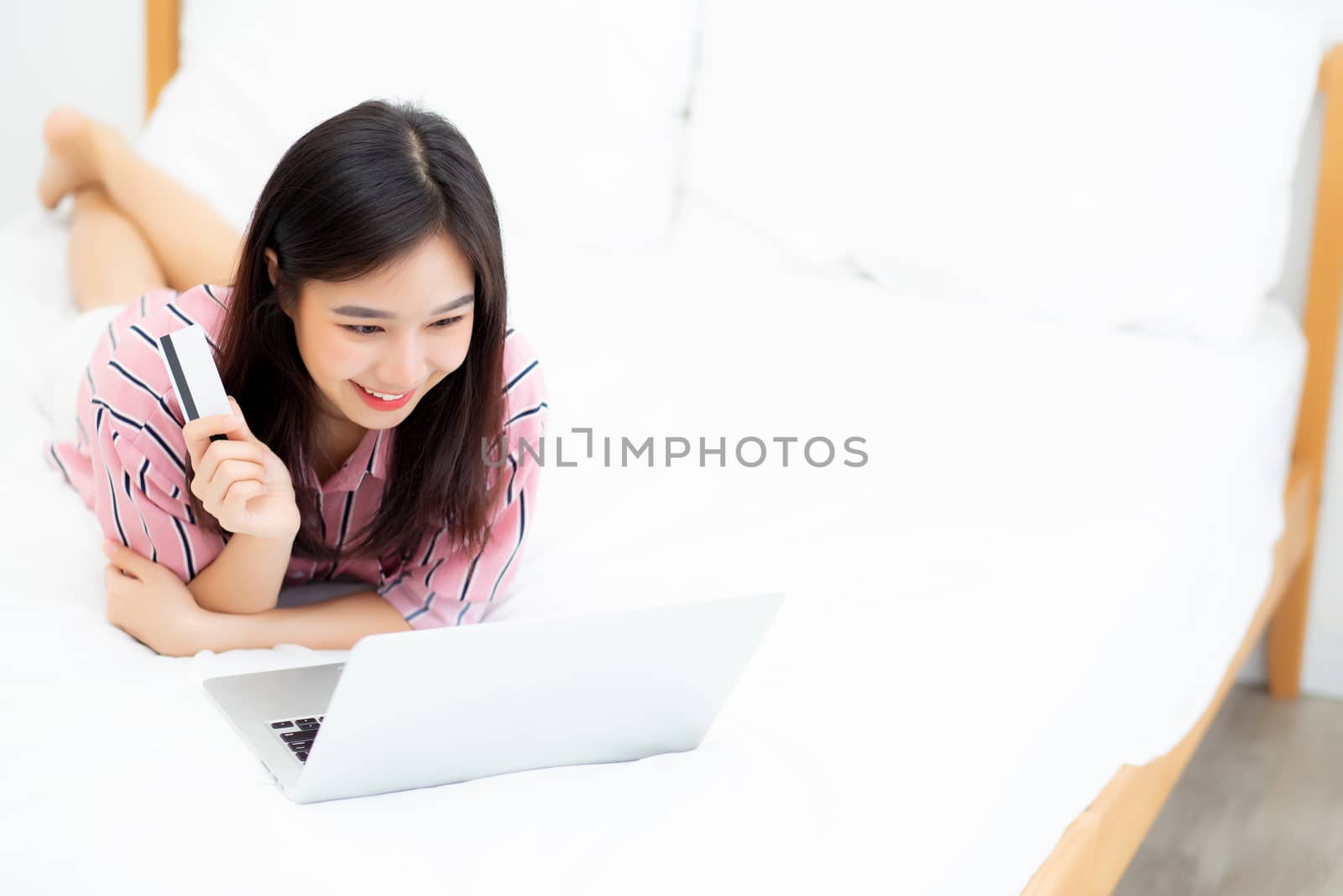 Beautiful of portrait young asian woman lying users credit card with laptop, content girl shopping online and payment with notebook computer on bed at bedroom, lifestyle concept.