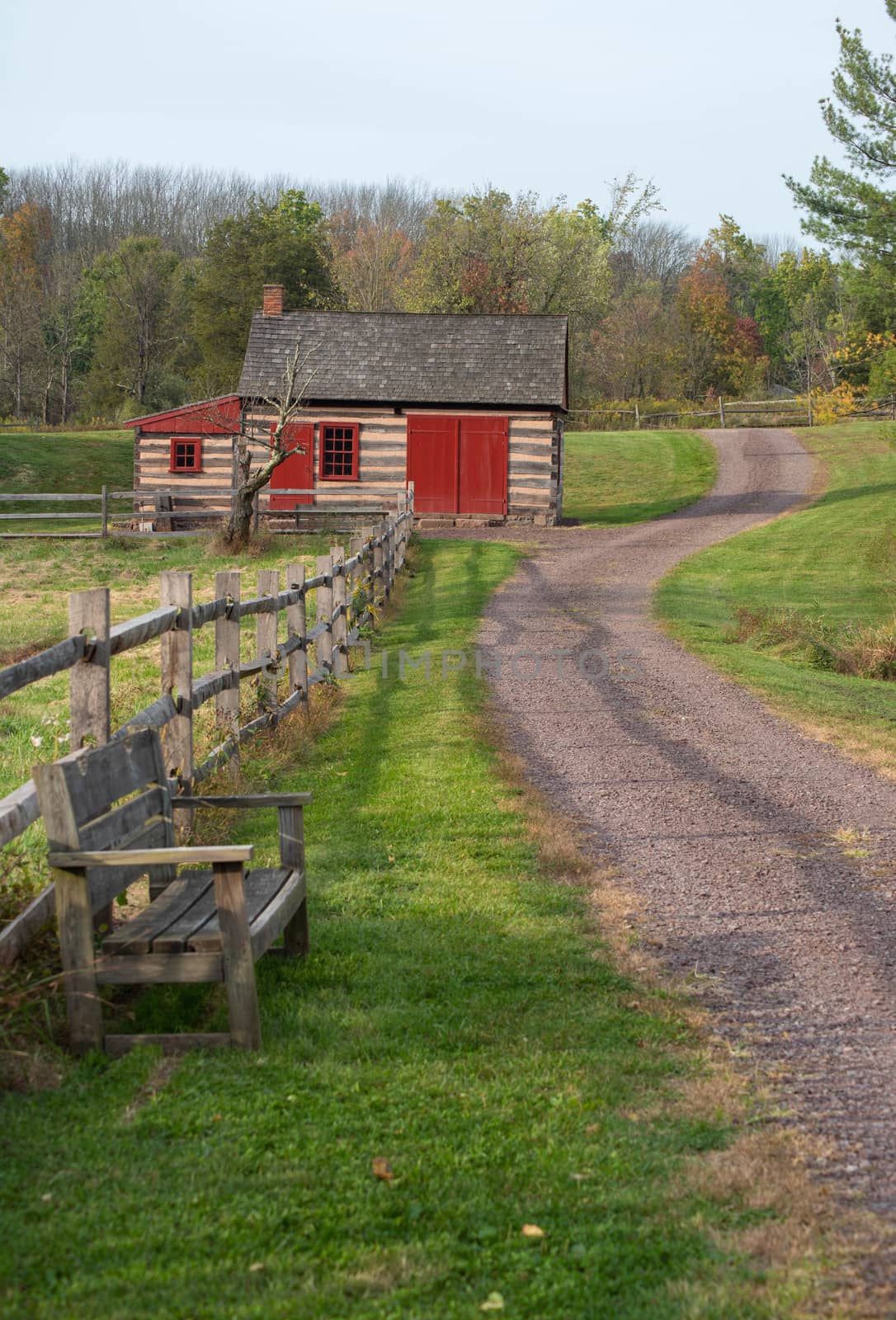 Rustic colonial Pennsylvania log cabin with red doors and windows along a winding footpath with an empty wooden bench. Green grass and Autumn foliage.