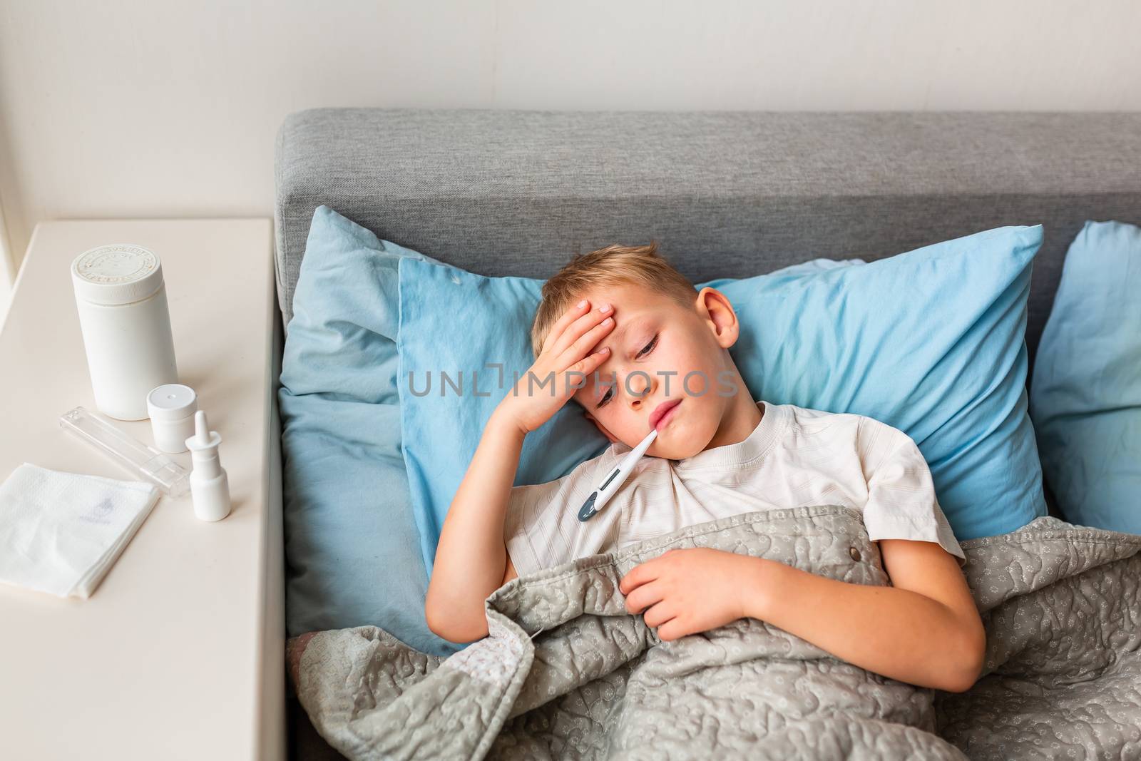 Sick little boy with high fever and headache laying in bed and holding thermometer in his mouth. Stay at home during corona virus epidemic if you feel sick