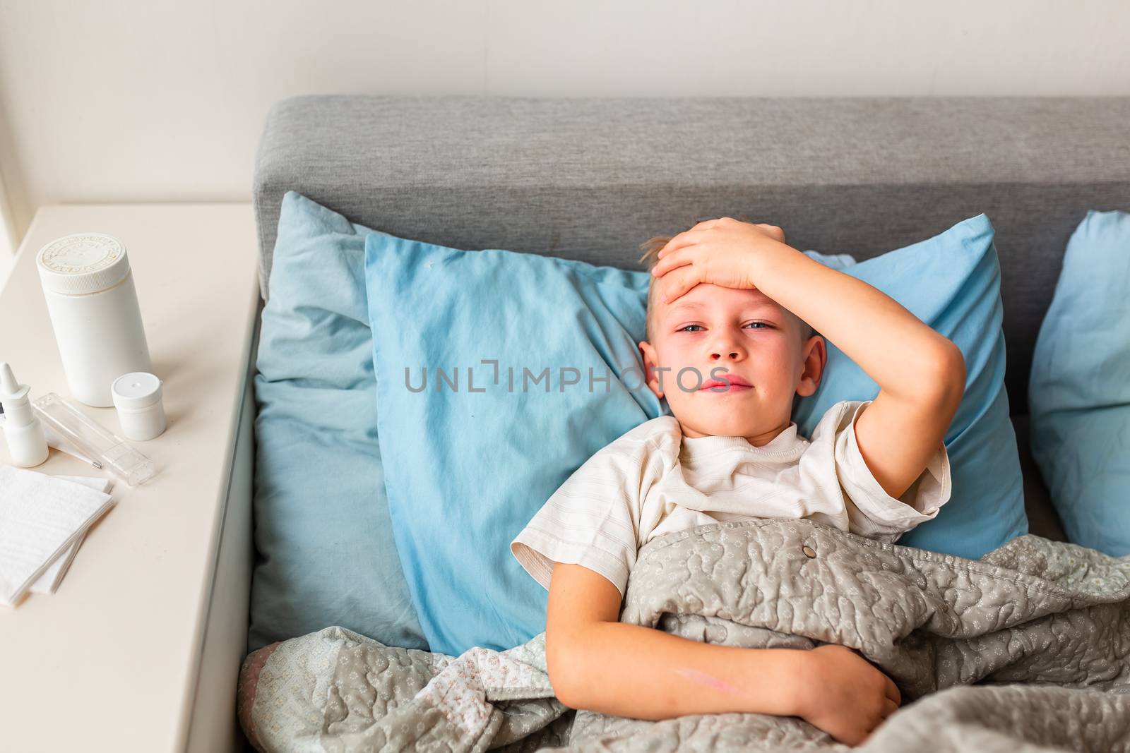 Sick little boy with high fever and headache laying in bed checking his forehead. Stay at home during corona virus epidemic if you feel sick