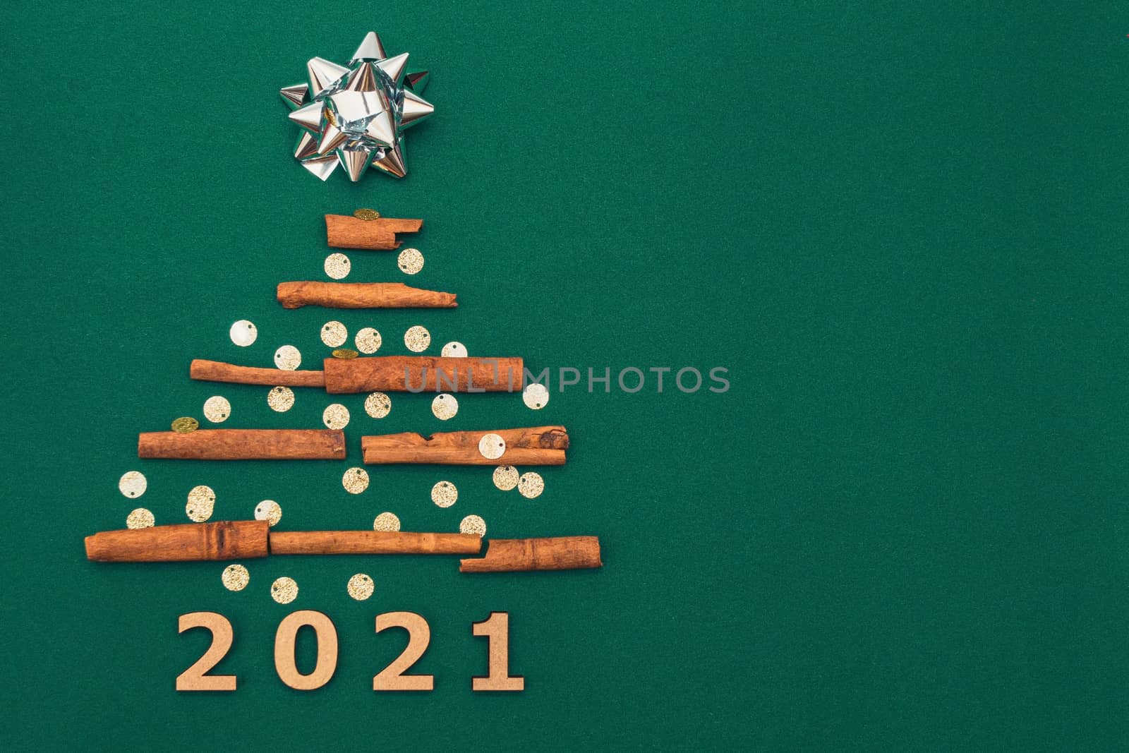 New Year card 2021. Christmas tree made of cinnamon sticks with silver gift bow on green background with silver confetti, copy space. Flat lay. template for design.