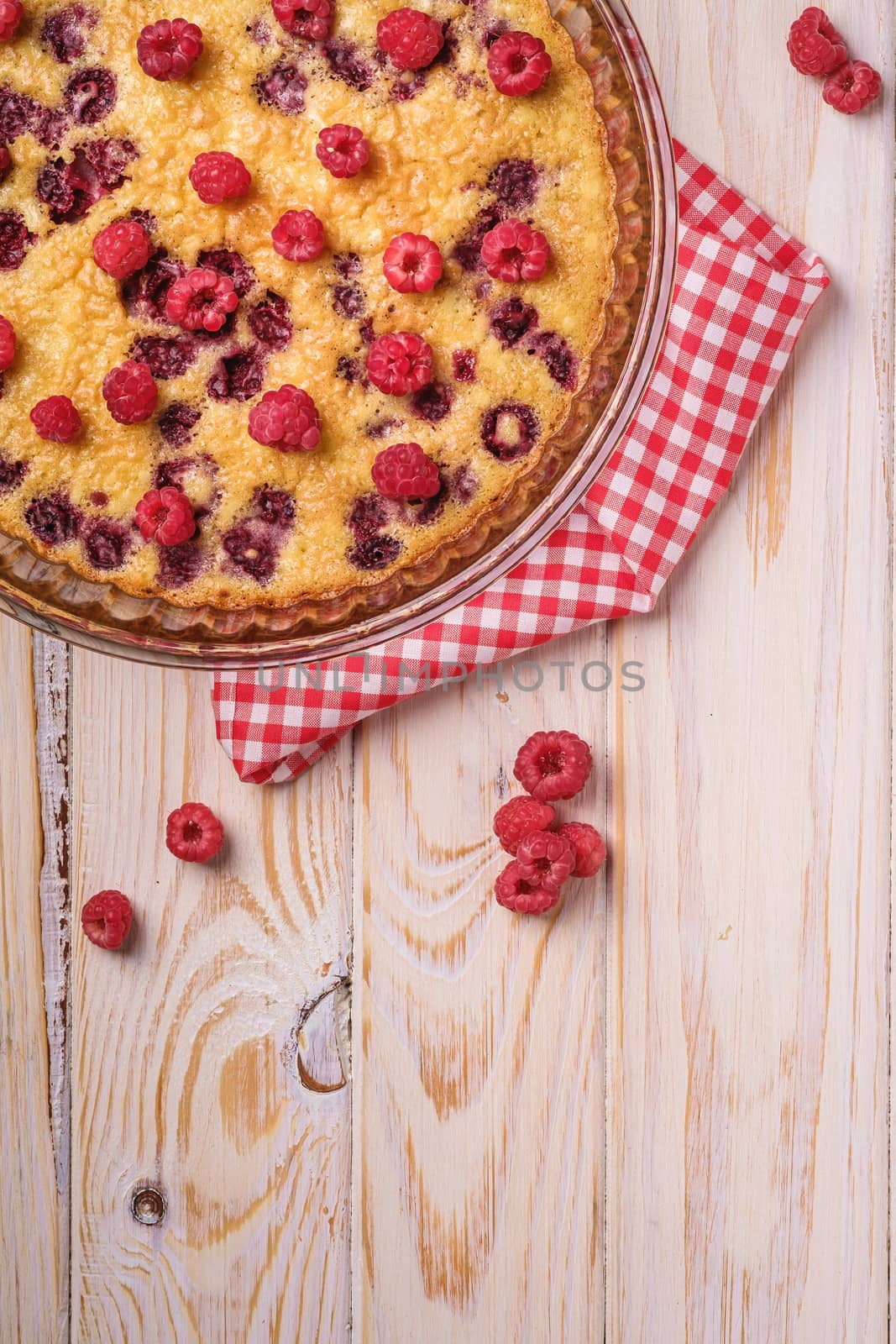 Sweet tasty pie with jellied and fresh raspberry fruits in baking dish with red tablecloth towel, wooden table background, top view