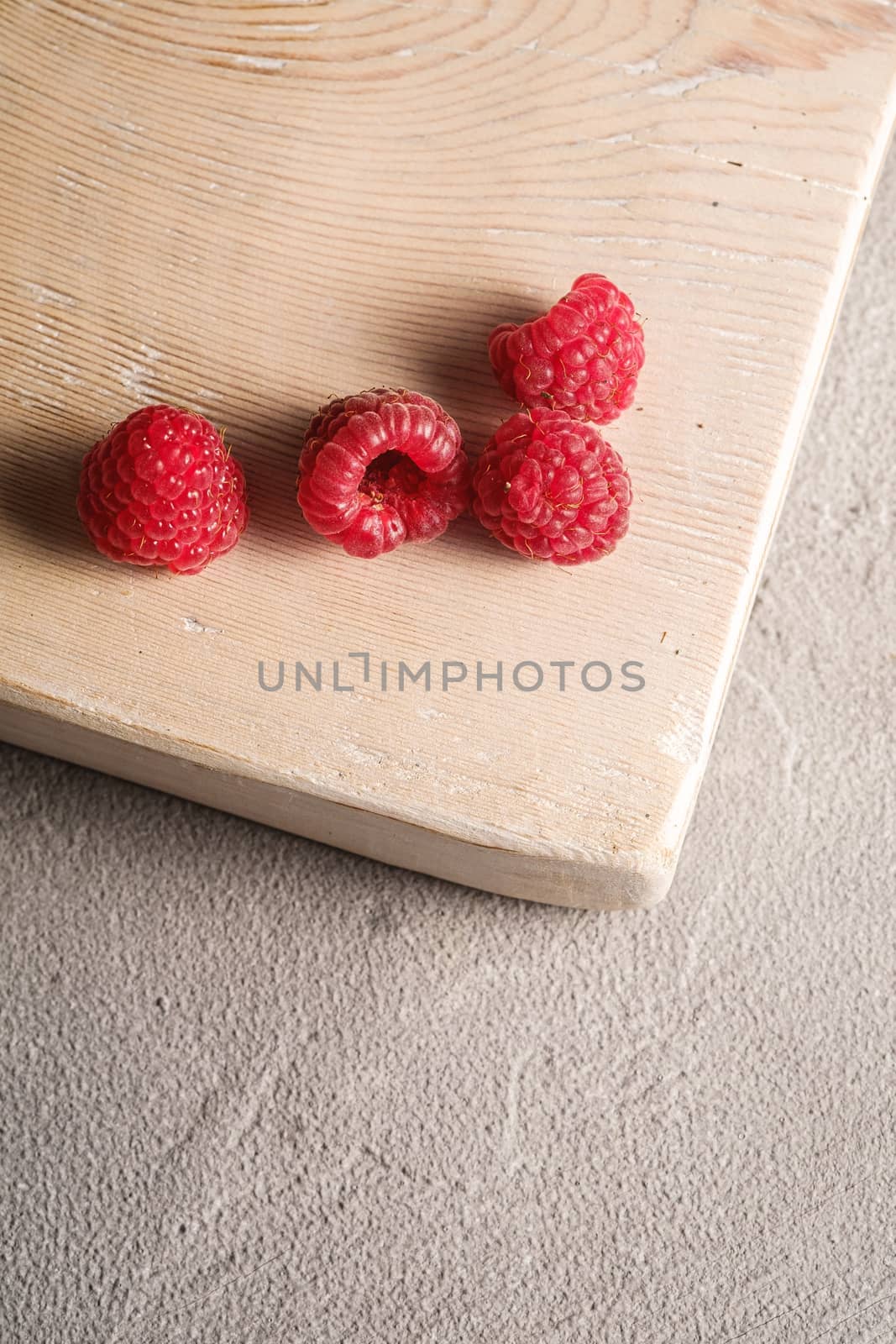 Raspberry fruits on old wooden cutting board, healthy pile of summer berries on stone concrete background, angle view