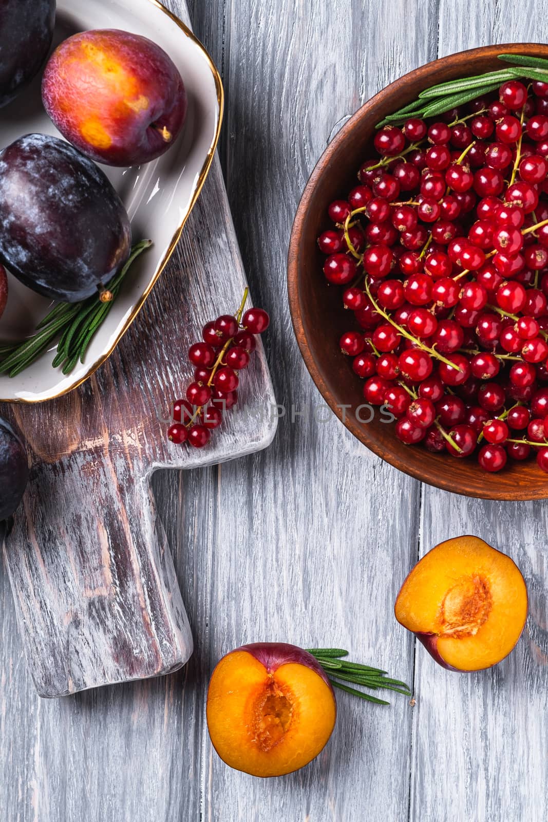 Fresh sweet plum fruits whole and sliced in plate with rosemary leaves on old cutting board with red currant berries in wooden bowl, grey wood background, top view