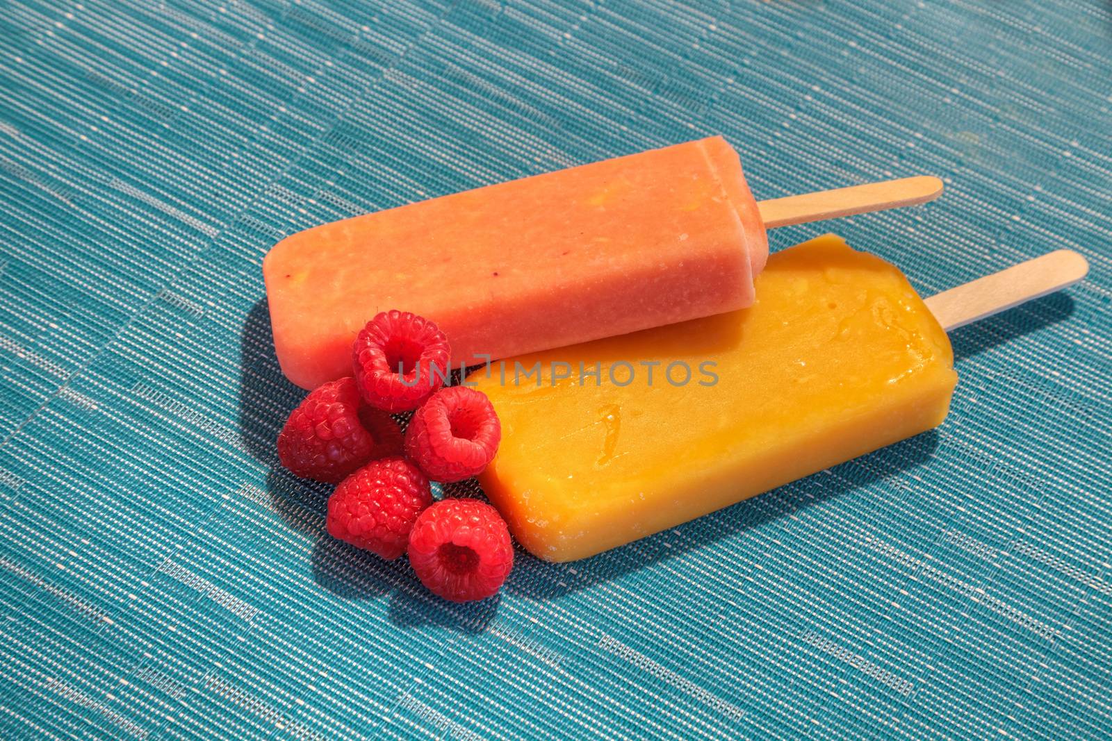 Fruit popsicles on an aqua blue background in summer including watermelon popsicle, tropical fruit popsicle, mango popsicle, and lime popsicle with fresh raspberries.