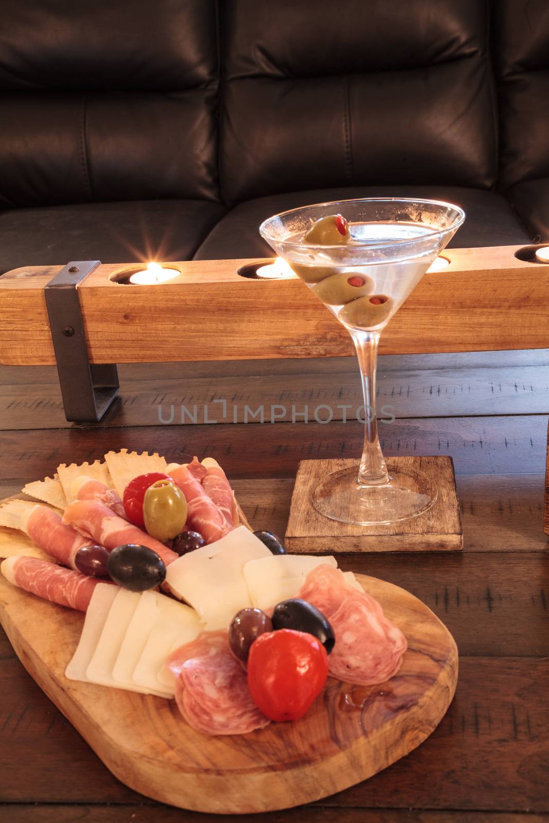 Martini with olives and a Charcuterie board on rustic wood with candles behind a spread of prosciutto panino, mozzarella cheese, Genoa salami, Fontina cheese and artisanal crackers.