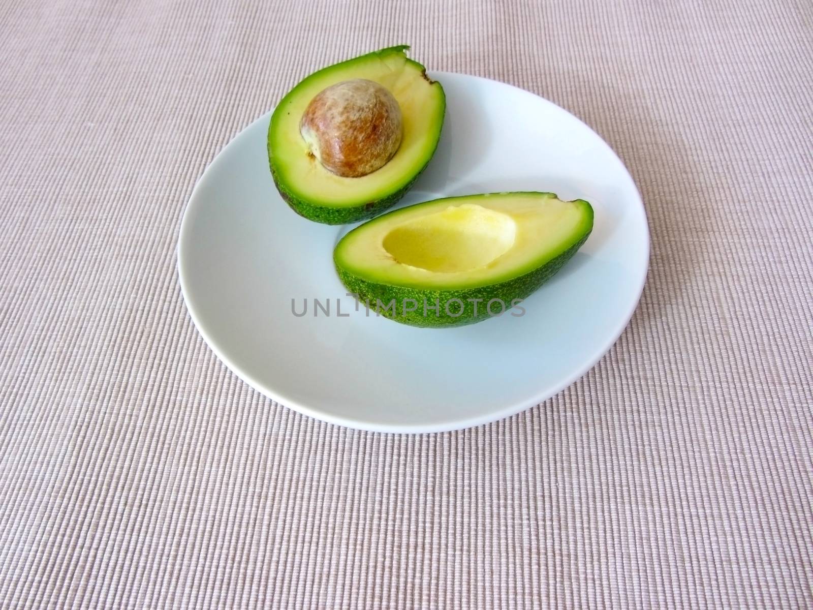 Fresh organic avocado halves, one with a bone, lie on a white plate by LanaLeta