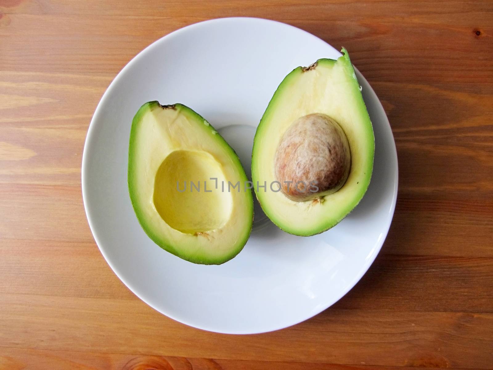 Close up fresh organic avocado halves, one with a bone, lie on a white plate on a wooden table, flat lay.