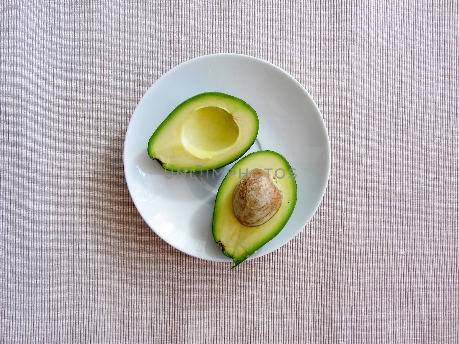Fresh organic avocado halves, one with a bone, lie on a white plate on a grey cotton striped textured canvas, flat lay. Healthy food concept..