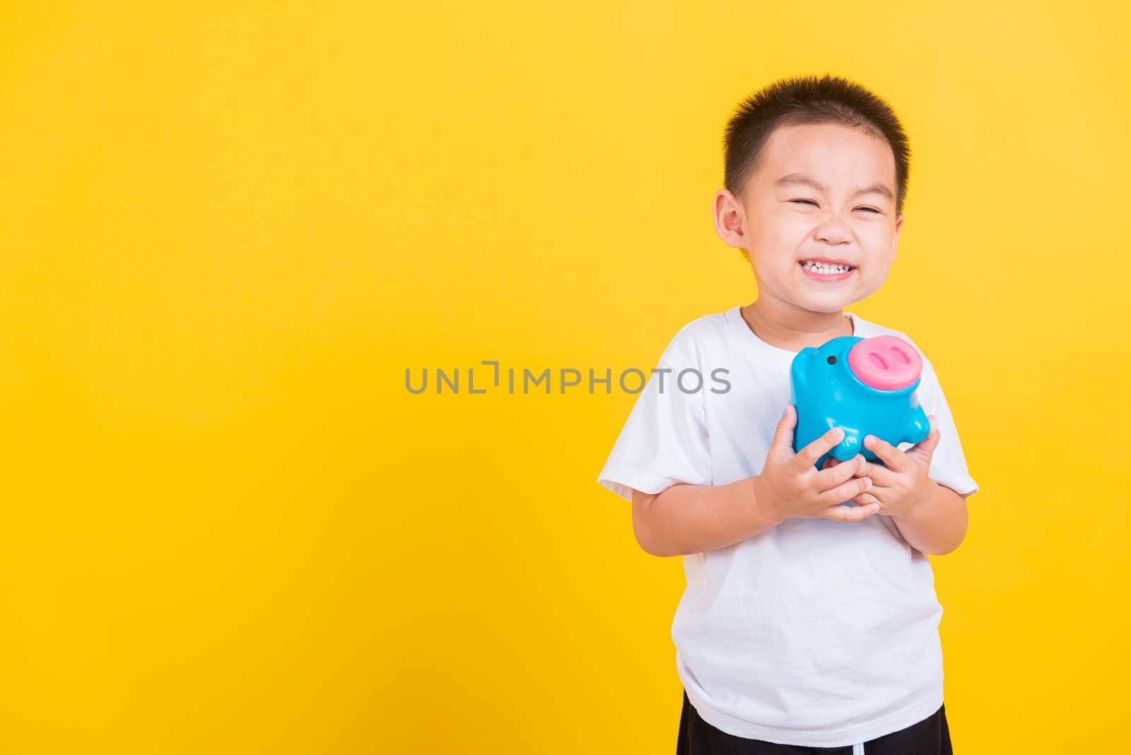 Asian Thai happy portrait cute little cheerful child boy smile holding piggy bank and looking camera, studio shot isolated on yellow background with copy space