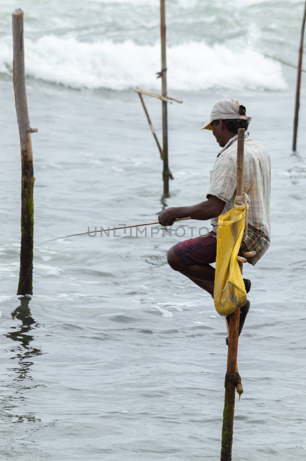 Stilt fisherman with his wooden rod facing side to the camera with his yellow pocket on the pole, fishing in a traditional unique method in Sri Lankan culture, sunny bright evening on the beach.