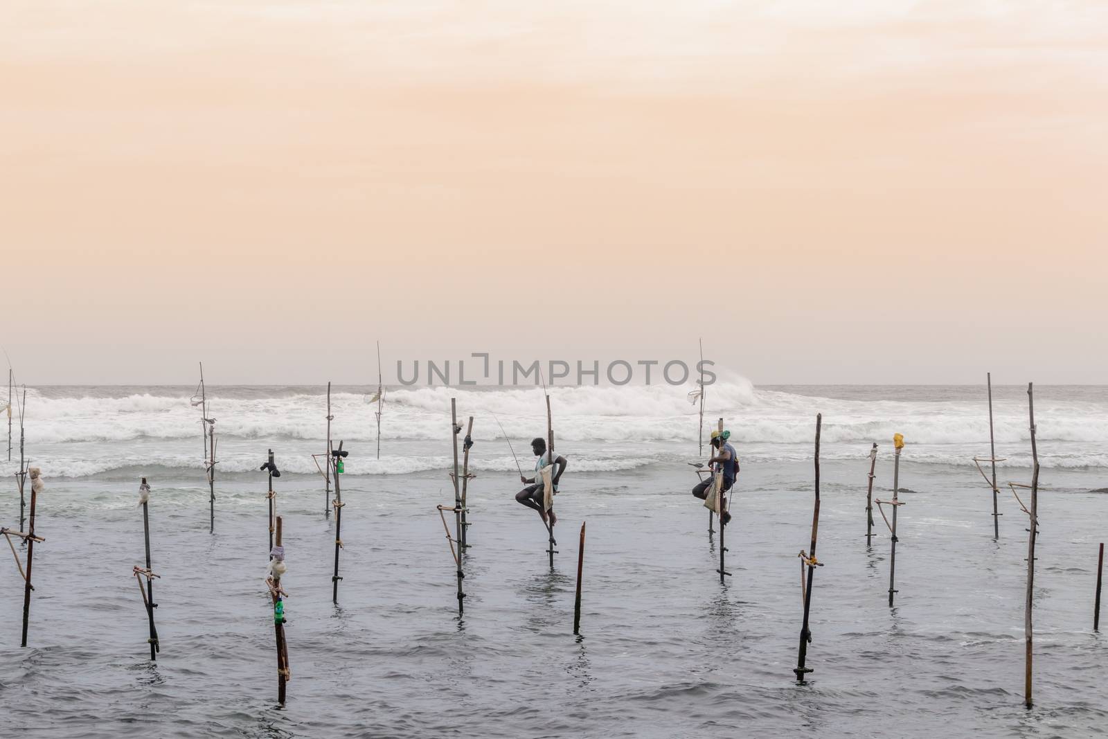 A couple of Stilt fishermen sitting on their poles with wooden fishing rods in their hands in the sunset evening. Ocean waves crash behind them in the background with an orange yellowish sky.A couple of Stilt fishermen sitting on their poles with wooden fishing rods in their hands in the sunset evening. Ocean waves crash behind them in the background with an orange yellowish sky.