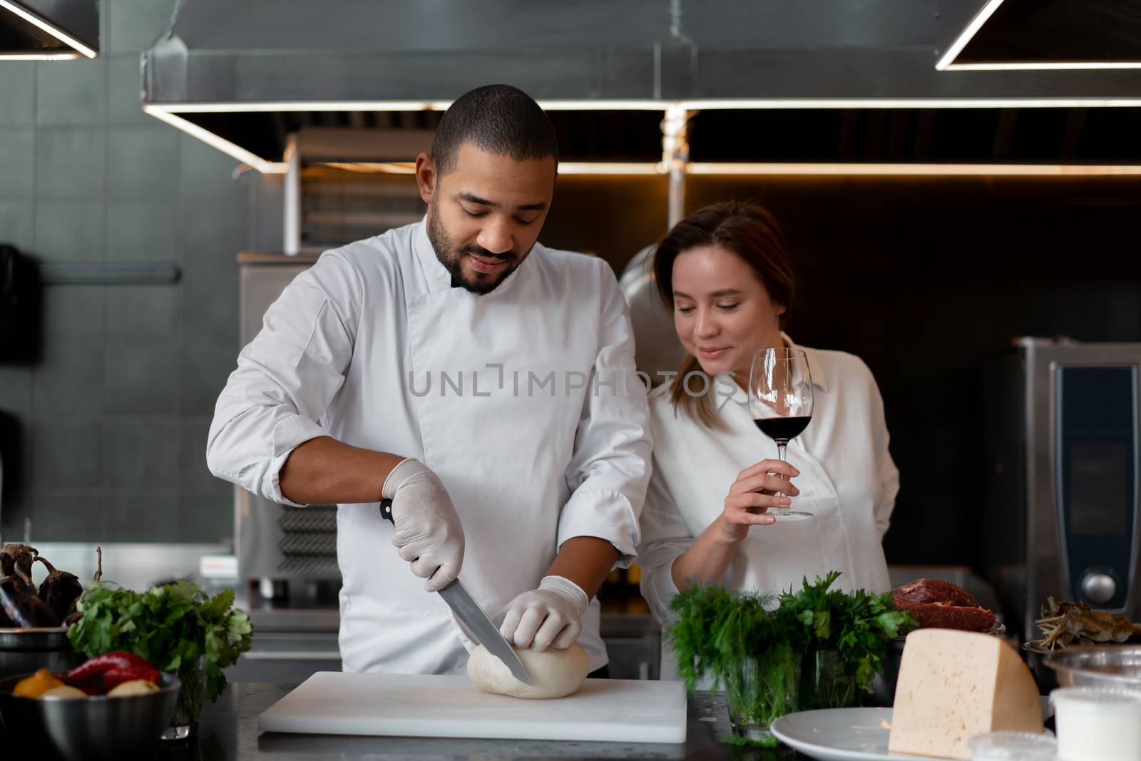 Handsome young African chef is cooking together with Caucasian girlfriend in the kitchen using red wine ingredient. A cook teaches a girl how to cook. Man and woman cooking in professional kitchen.