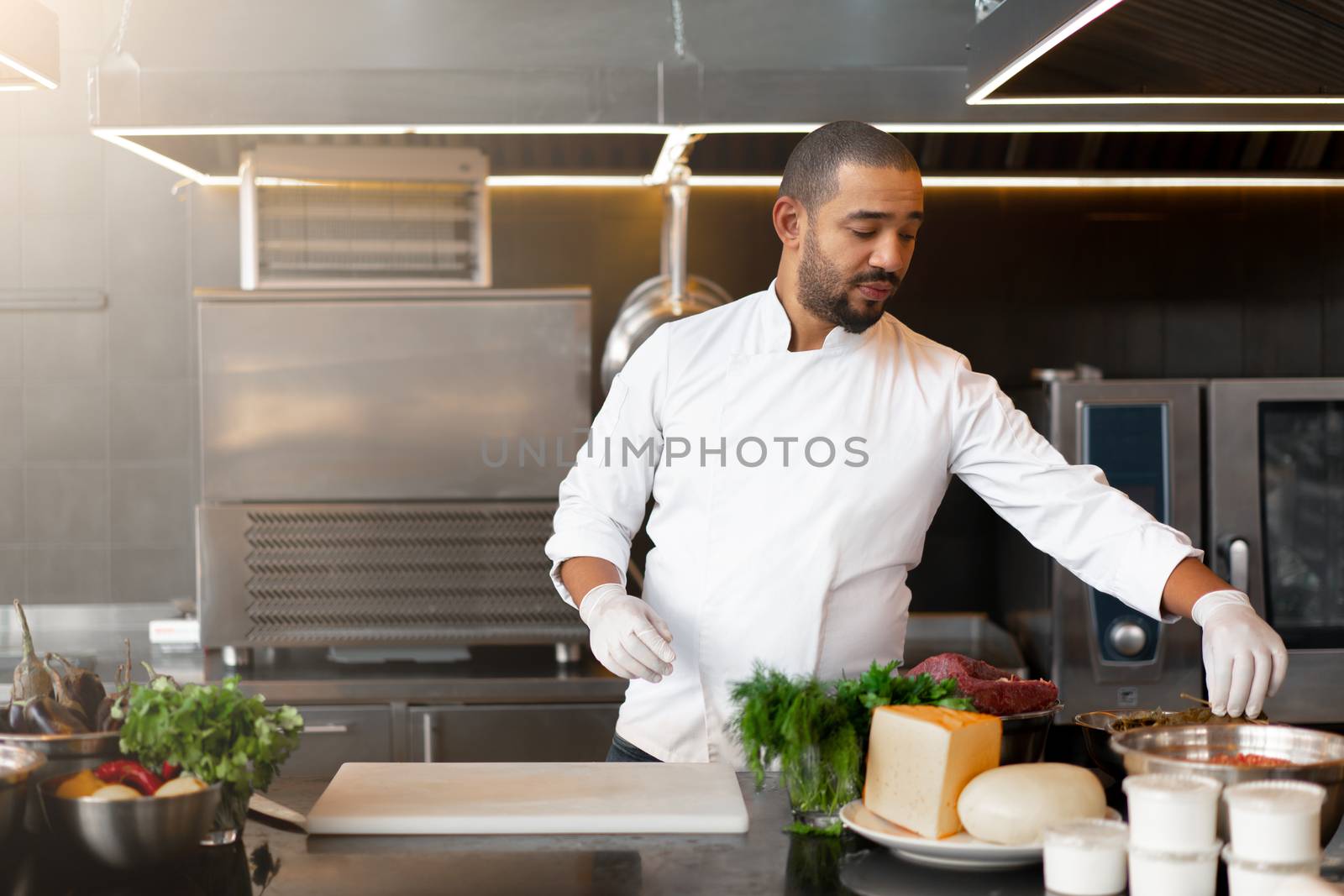 Handsome young African chef standing in professional kitchen in restaurant preparing a meal of meat and cheese vegetables. Portrait of man in cook uniform. Healthy eating concept.