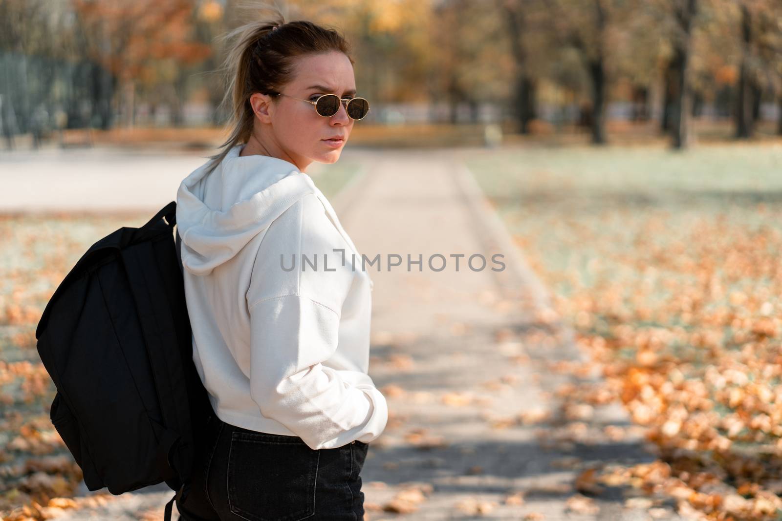 A young beautiful woman with a ponytail and sunglasses, with a backpack on his shoulders in the park. Photo from the back. White hoodie