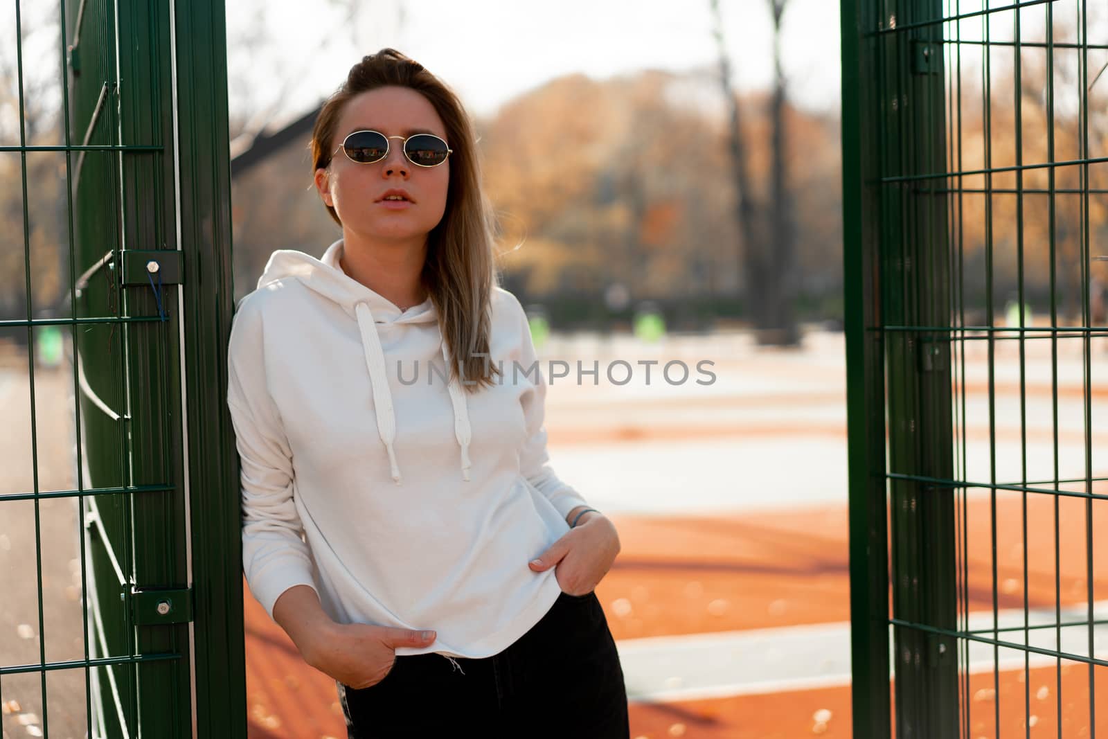 Outdoor close up portrait of young beautiful woman with long hair in sunglasses, dressed in a white hoodie sweater, near the sportsground. youth culture summer pastime