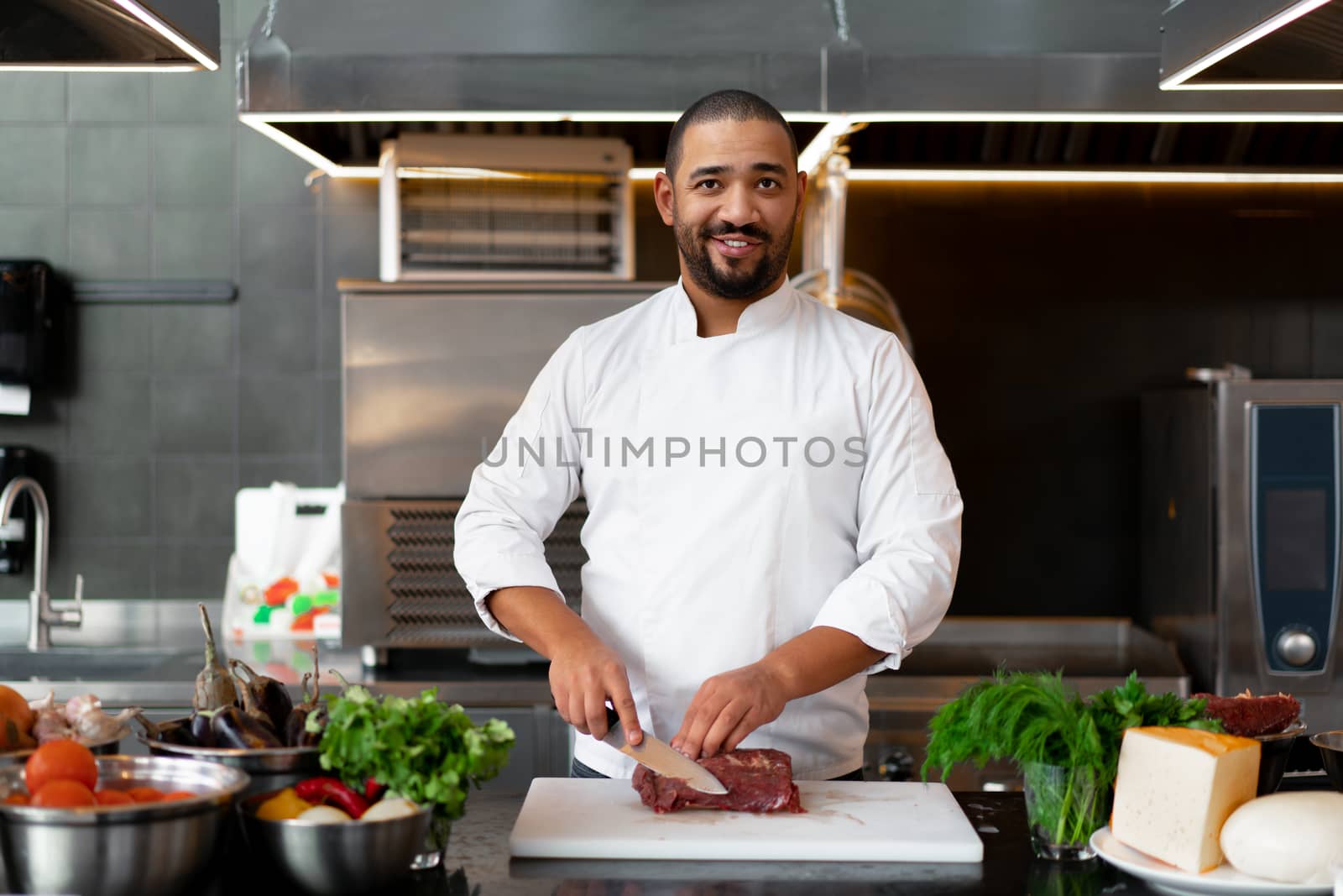 Handsome young African chef standing in professional kitchen in restaurant preparing a meal of meat and cheese vegetables. Portrait of man in cook uniform Cuts meat with a metal knife.