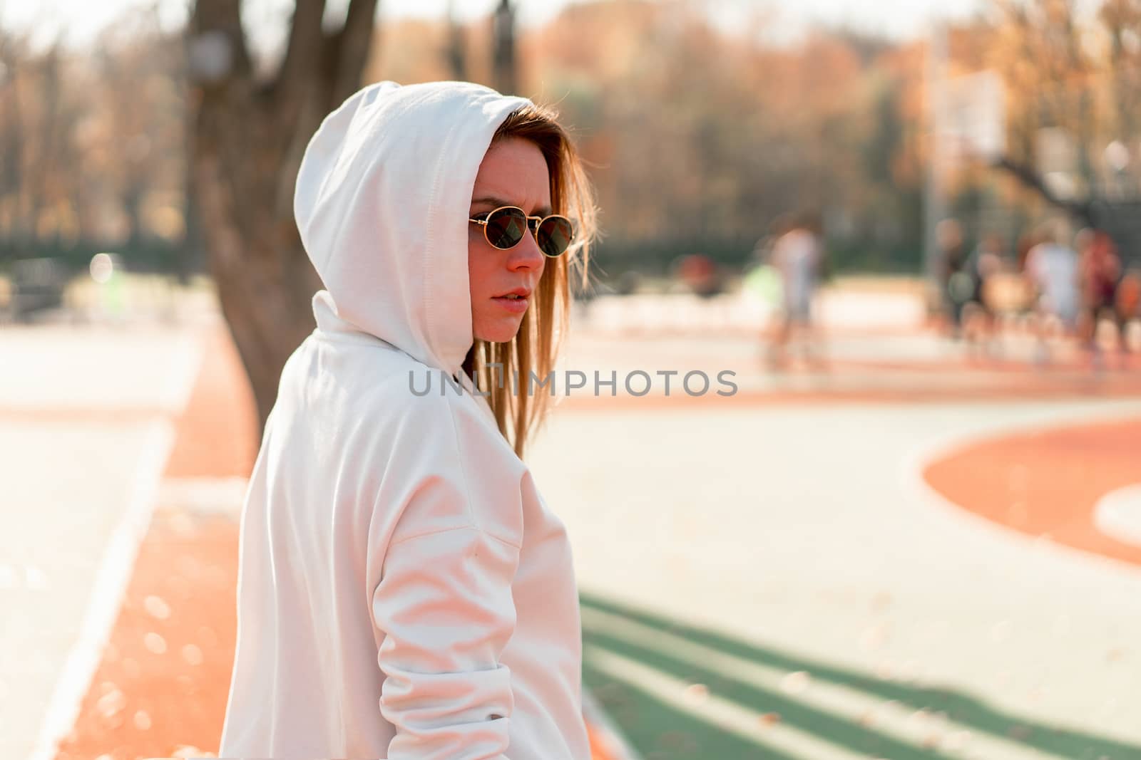 Outdoor close up portrait of young beautiful woman with in glasses, dressed in a white sweater, near the sportsground. Woman in a hood turned her head to the camera. White hoodie