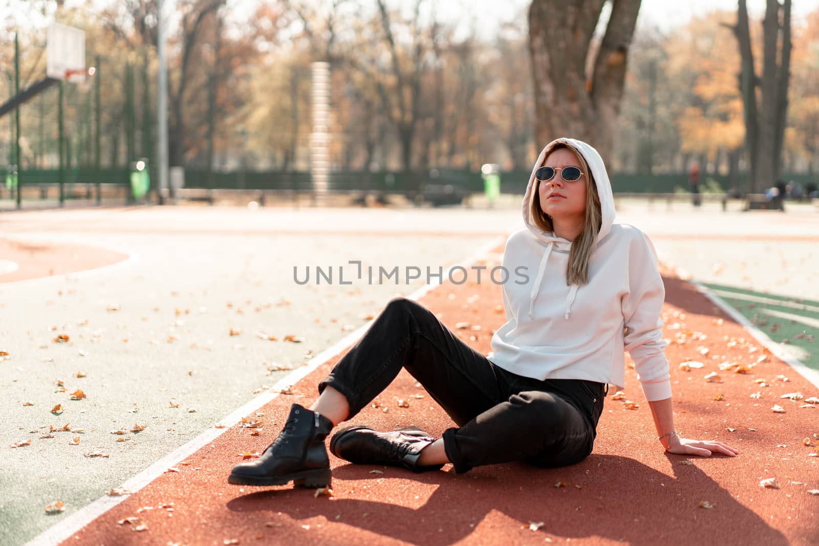 Outdoor portrait of young beautiful woman with long in sunglasses and a white hooded sweater sitting on the sportsground track. youth culture summer pastime
