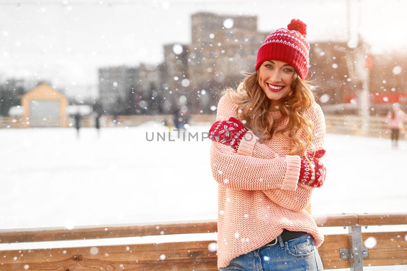Beautiful lovely middle-aged girl curly hair warm peach sweater red knitted hat glove stands ice rink background Town Square Christmas mood lifestyle Happy holiday woman snowy day Winter leisure