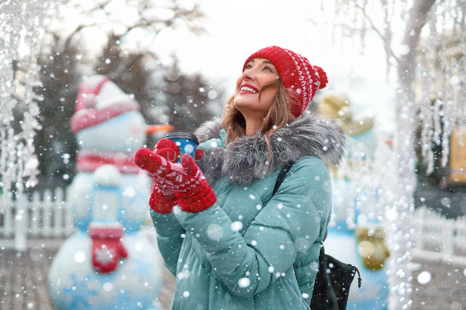 Beautiful lovely middle-aged girl curly hair warm winter jackets red knitted hat glove stands ice rink background Town Square Christmas mood lifestyle Happy holiday woman drink hot beverage coffee