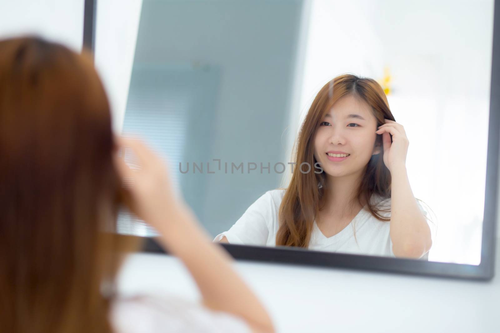 Beautiful of portrait asian young woman examining with face and smile looking on mirror at bedroom, girl beauty of makeup and dressed up with reflection, lifestyle and wellness concept.