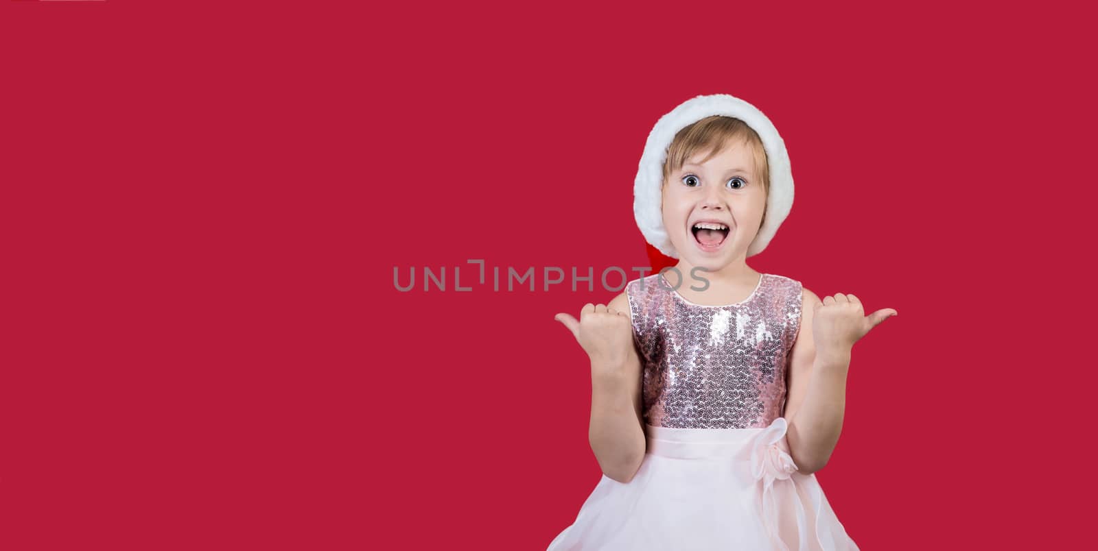 Surprised funny cacusian cute child girl in santa hat looking at the camera, shouting happily and showing thumbs up isolated on red background. Merry Christmas presents shopping sale..