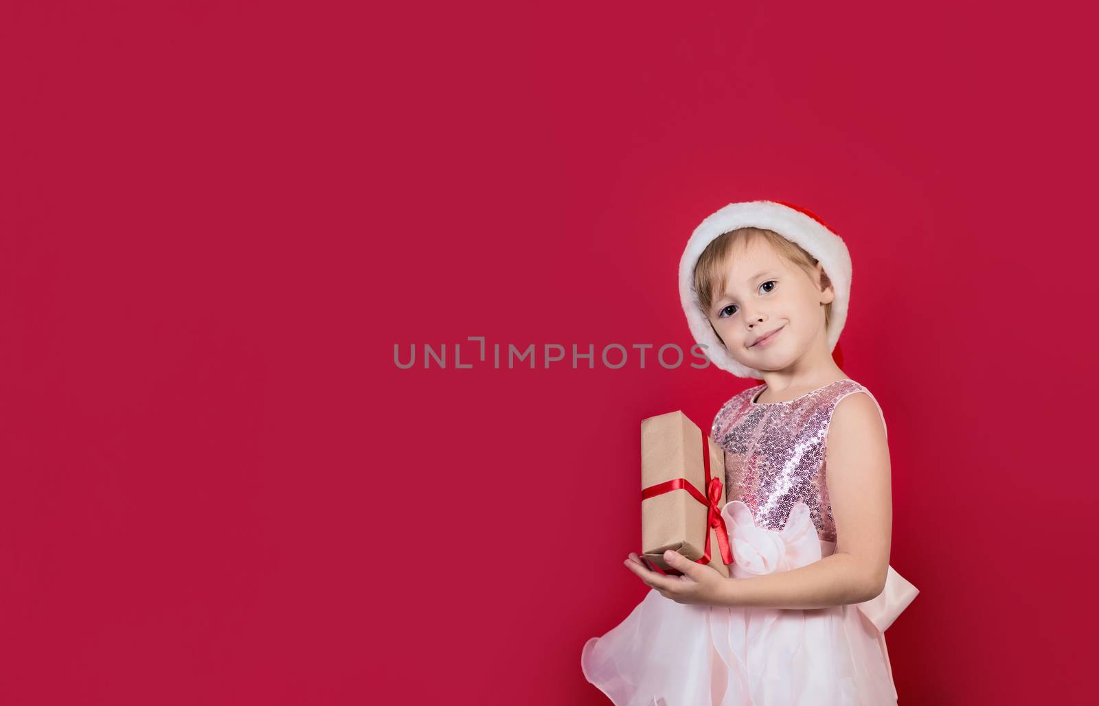 Smiling funny cacusian cute child girl in santa hat looking at the camera with gift box celebrating happy 2021 New Year isolated on red background. Merry Christmas presents shopping sale.