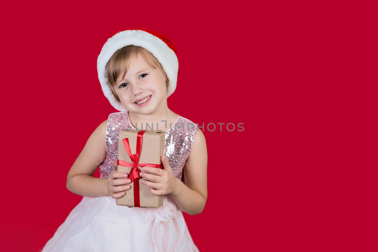 Surprised funny cacusian cute child girl in santa hat looking at the camera, smiling widly happily and hugs a gift isolated on red background. Merry Christmas presents shopping sale.