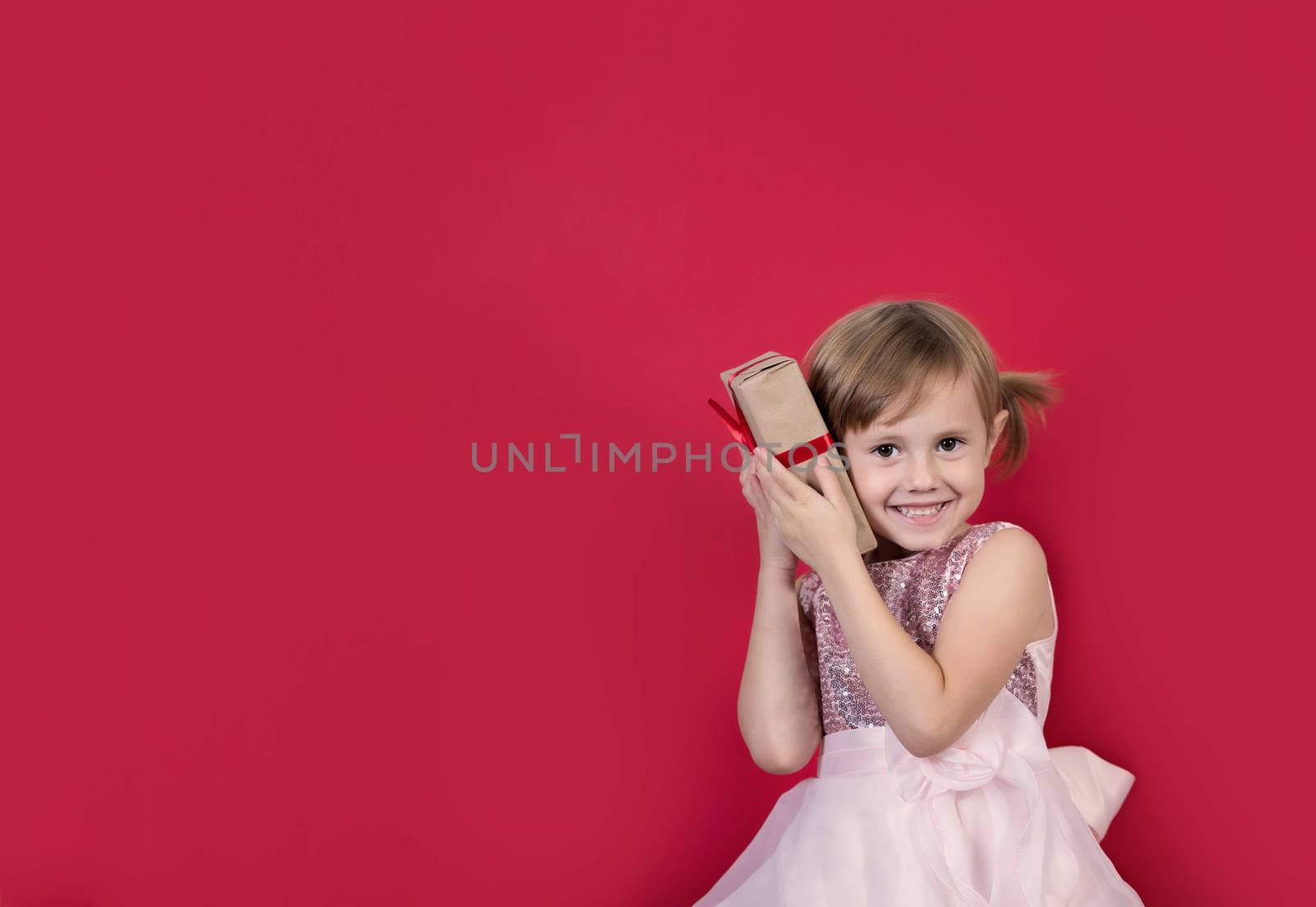 Smiling cute cacusian little girl in santa hat looking at the camera with gift box near her ear celebrating happy 2021 New Year isolated on red background. Merry Christmas presents shopping sale.