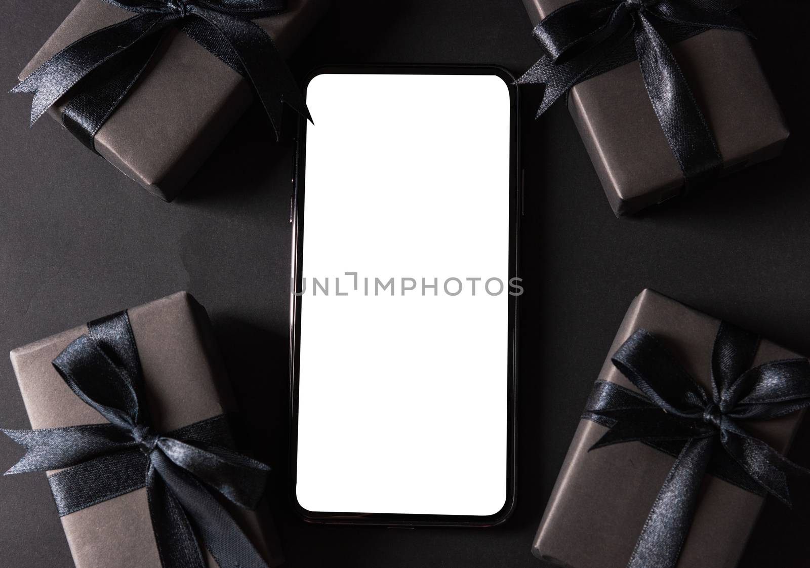 Black Friday sale shopping concept, Top view of gift box wrapped black paper and black bow ribbon present around smartphone blank screen, studio shot on dark background