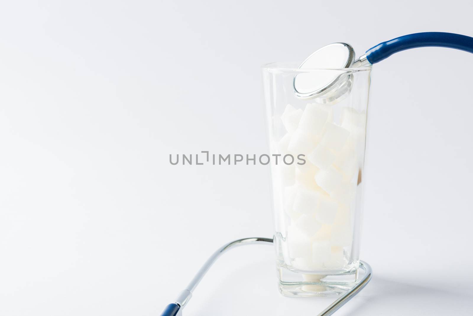 A glass full of white sugar cube sweet food ingredient and doctor stethoscope, studio shot isolated white background, health high blood risk of diabetes and calorie intake concept and unhealthy drink