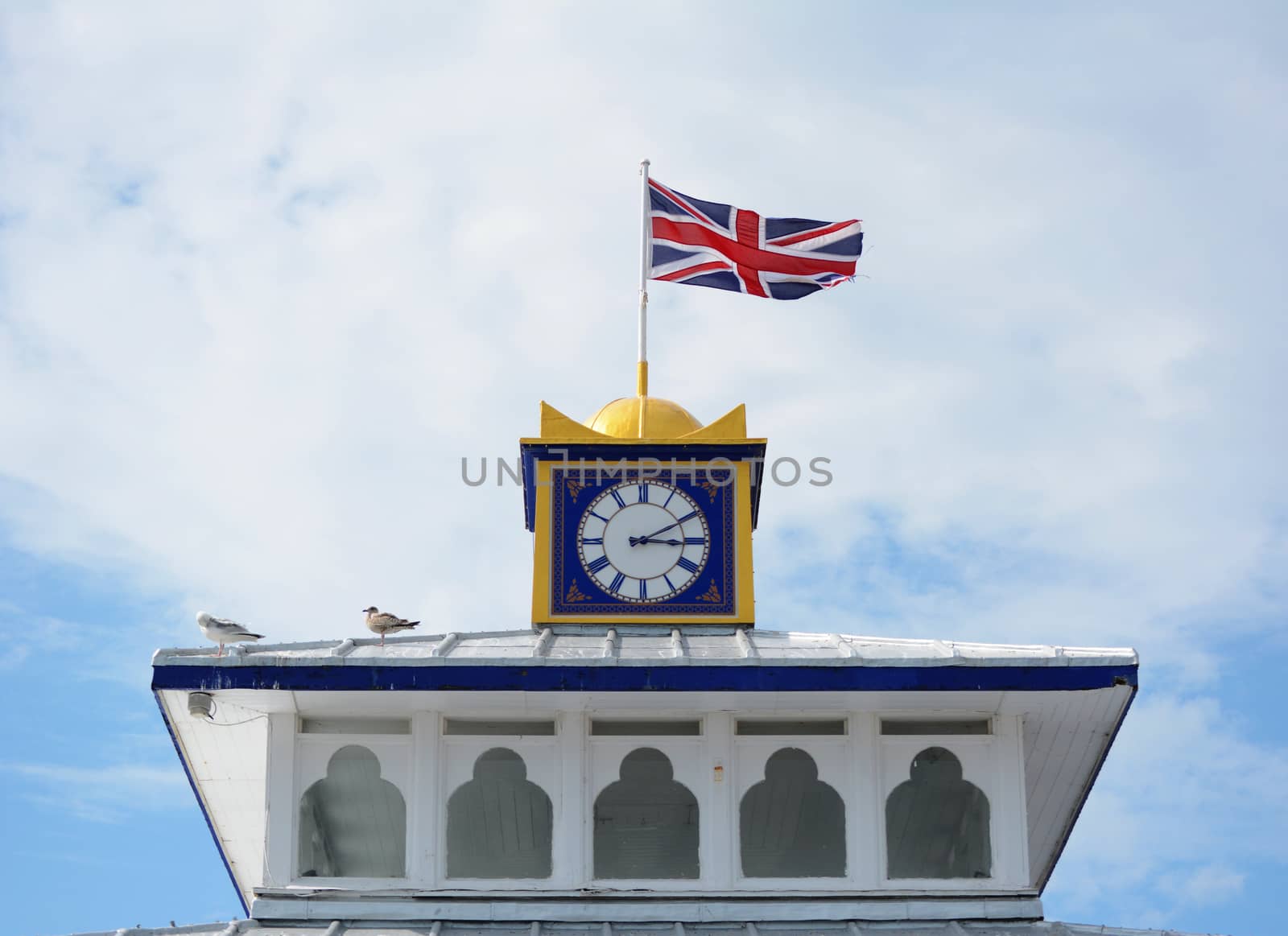 The Union Jack flag flies atop the clock tower of Eastbourne pier. Gulls sit on the roof in sunshine.