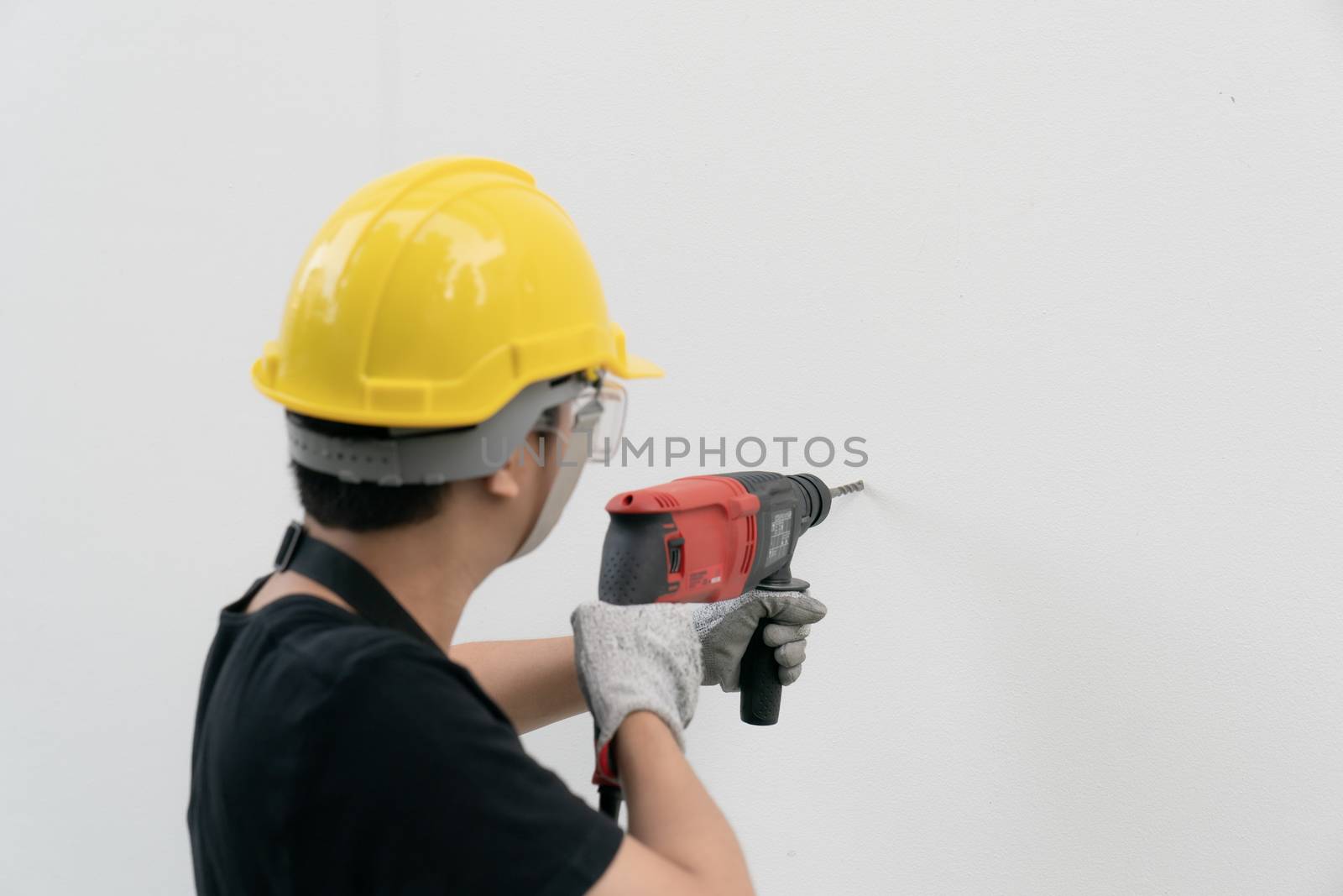 Man builder or worker with yellow helmet and goggle drilling machine on white wall.