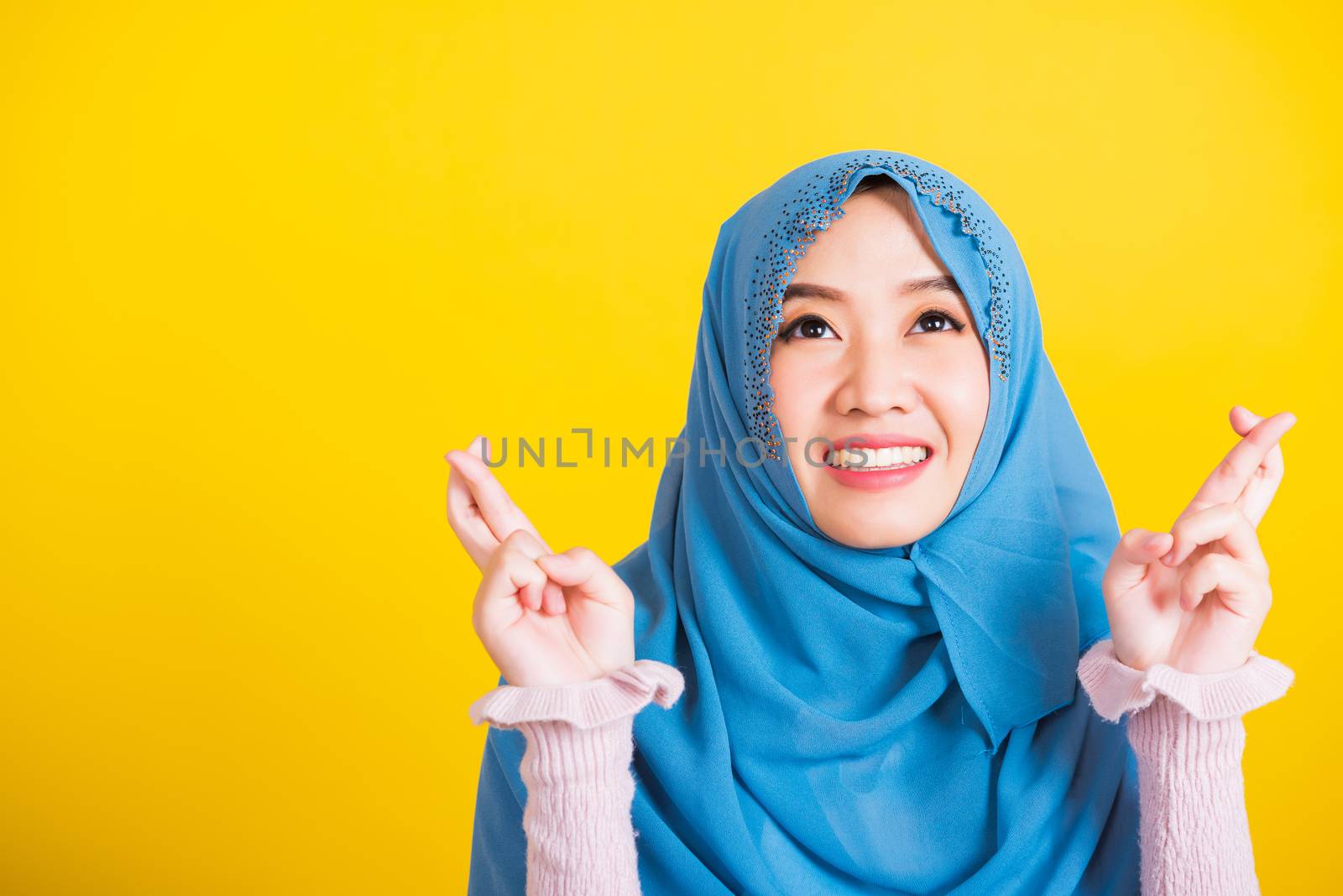 Asian Muslim Arab, Portrait of happy Asian beautiful young woman Islamic religious wear veil hijab she smiling and holding fingers crossed for good luck, studio shot isolated on yellow background