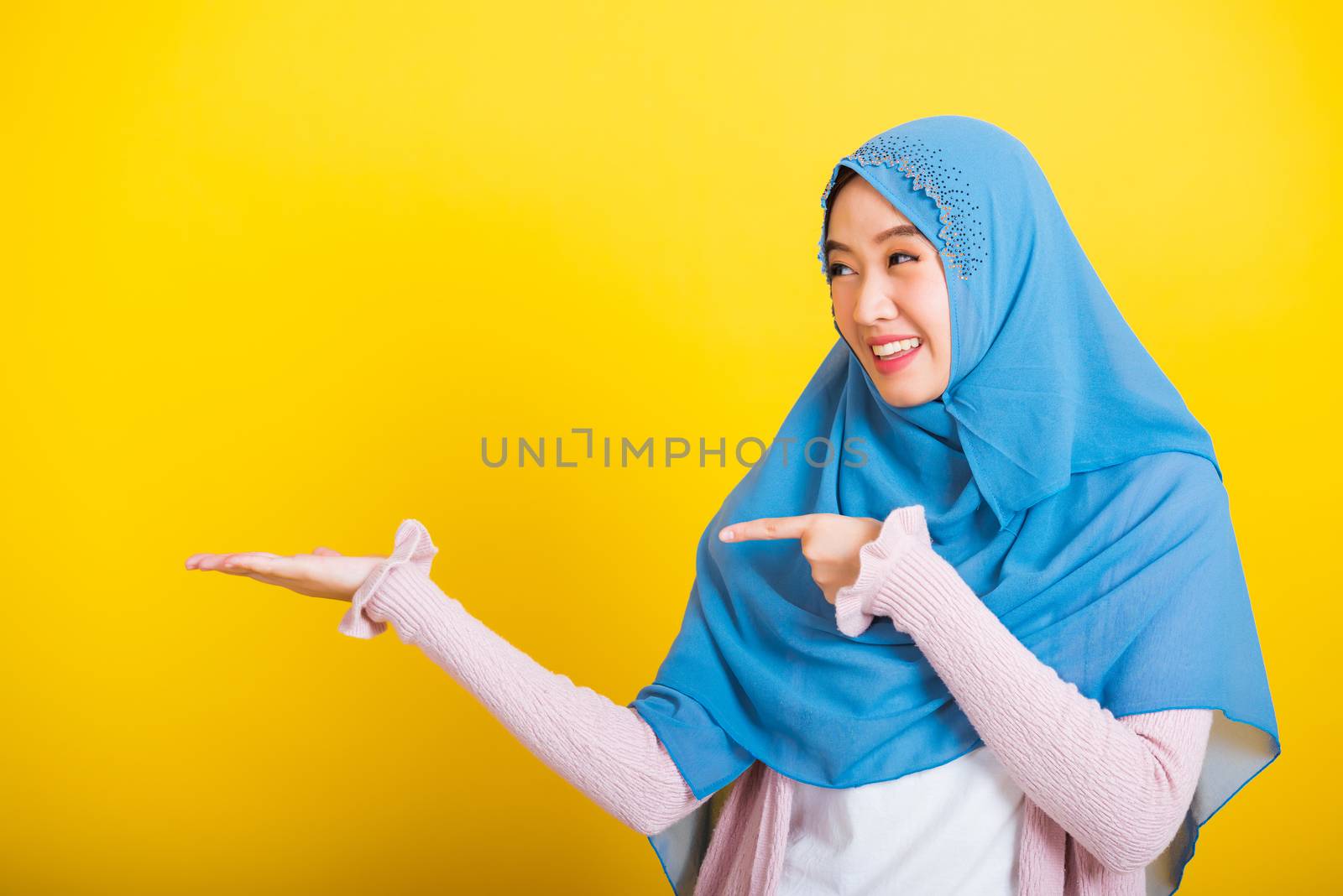 Asian Muslim Arab, Portrait of happy beautiful young woman Islam religious wear veil hijab funny smile she positive expression pointing finger side sideways to space isolated yellow background