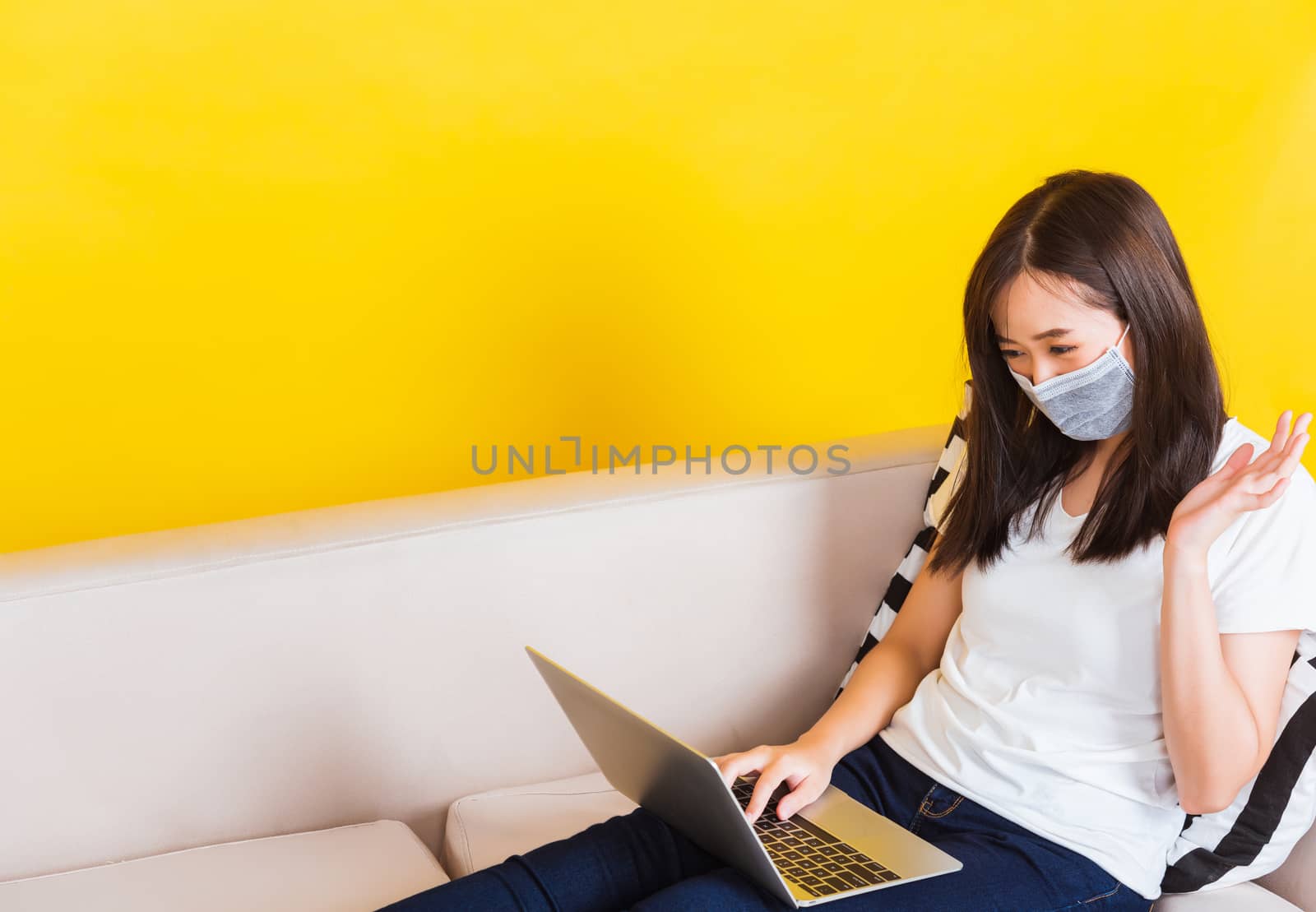 Portrait Asian of beautiful young woman sitting on sofa wearing medical face mask protective she work from home with laptop computer during Coronavirus studio shot isolated on yellow background