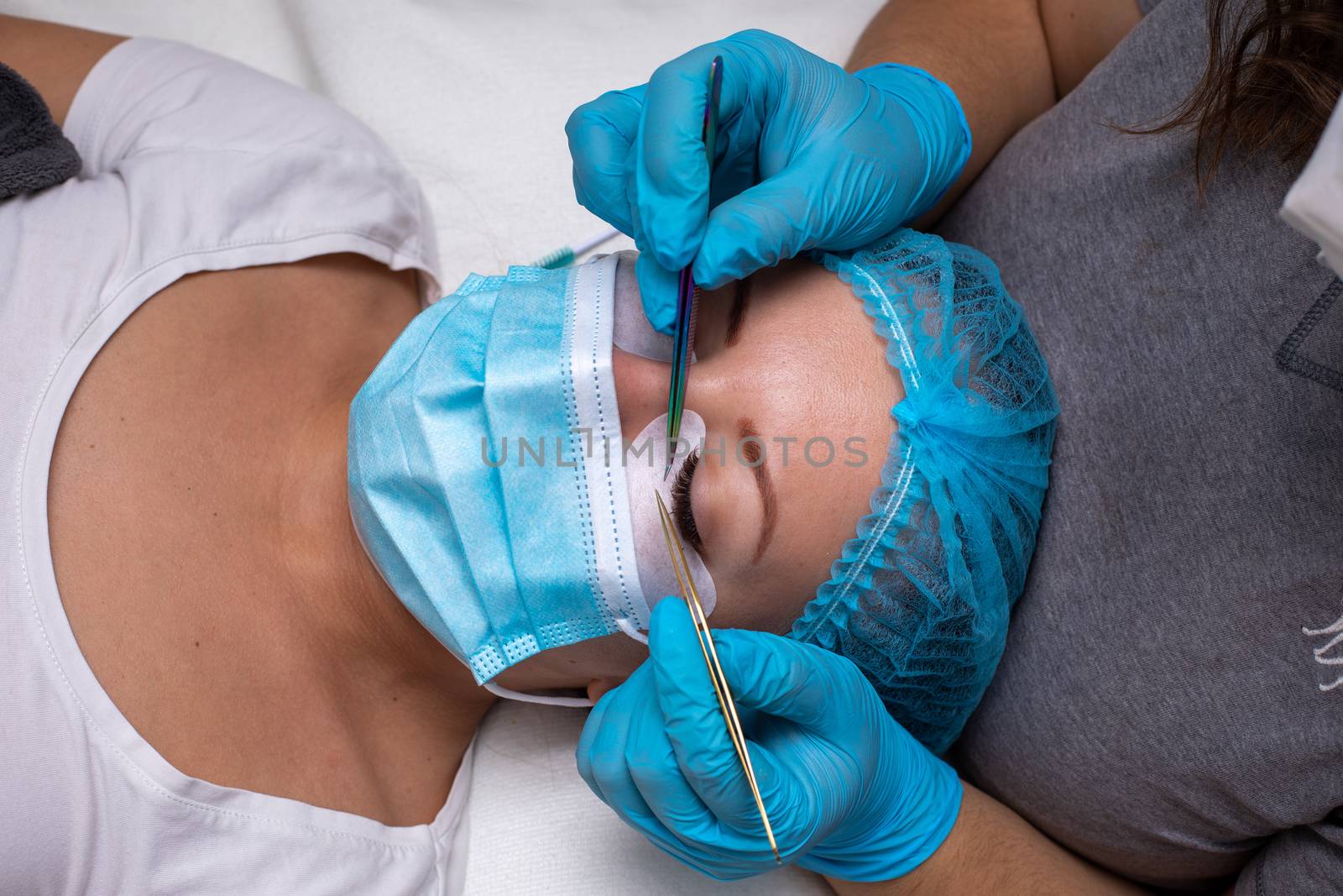 Treatment beauty procedure of upgrade Eyelashes Extension during a pandemic with preventive measures. Woman face wirh protective cap and facemask