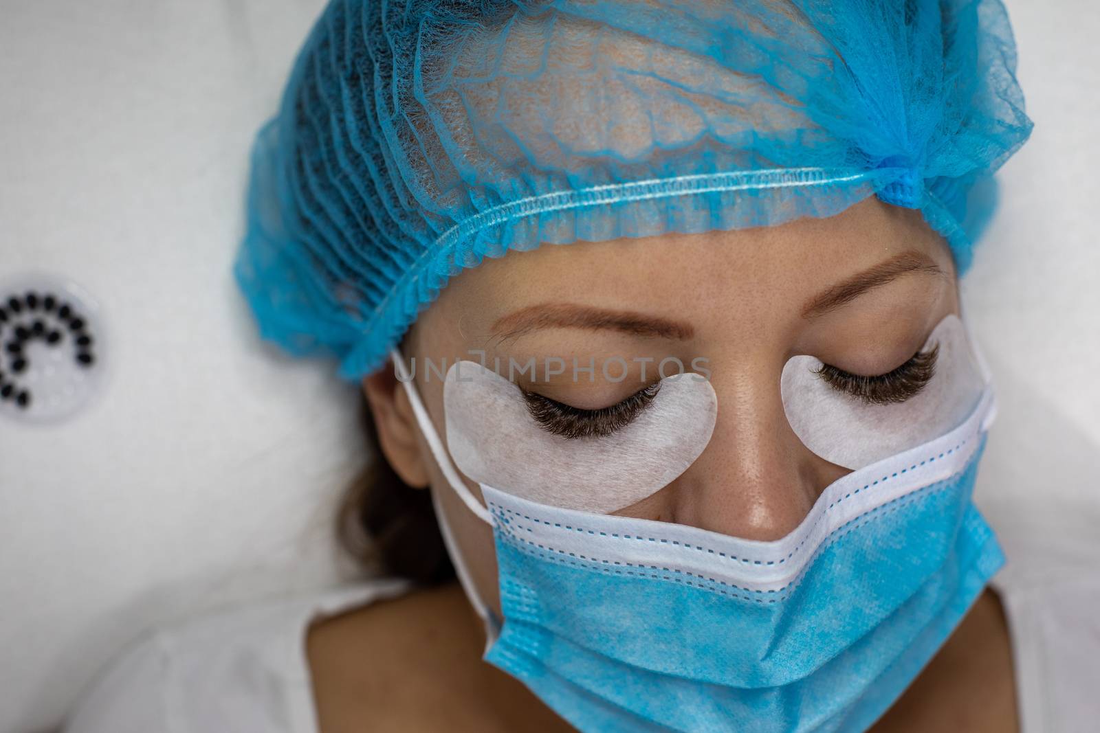 Treatment beauty procedure of upgrade Eyelashes Extension during a pandemic with preventive measures. Woman face wirh protective cap and facemask