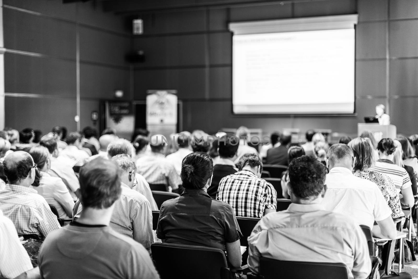 Female speaker giving presentation in lecture hall at university workshop. Audience in conference hall. Rear view of unrecognized participant in audience. Scientific conference event.