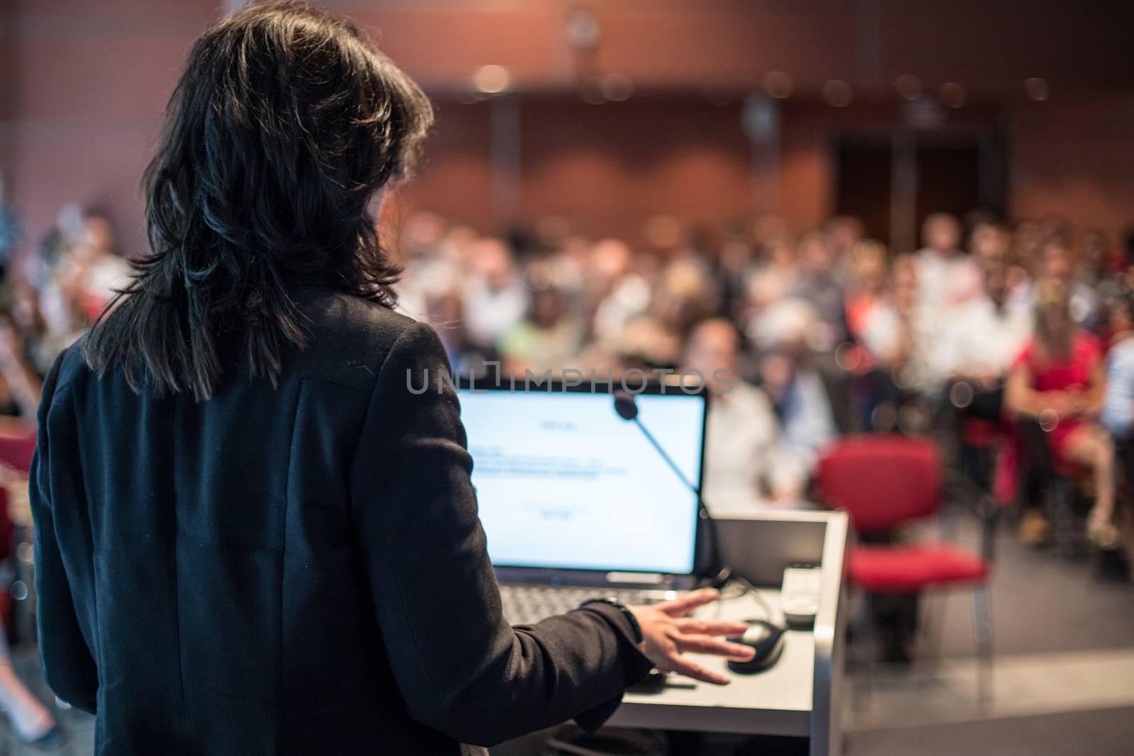 Female speaker giving a talk on corporate business conference. Unrecognizable people in audience at conference hall. Business and Entrepreneurship event.